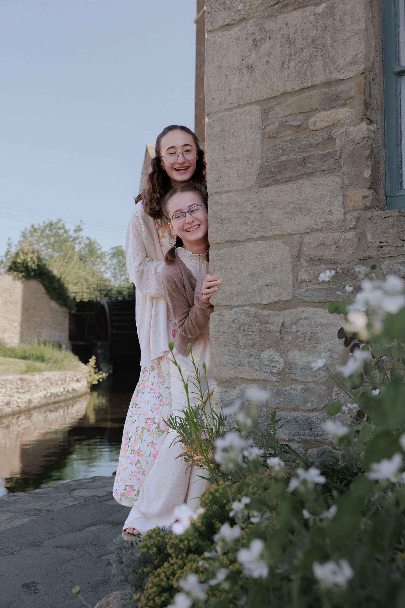 Two young women smiling and peeking around a stone wall beside a canal, surrounded by white wildflowers, creating memories.