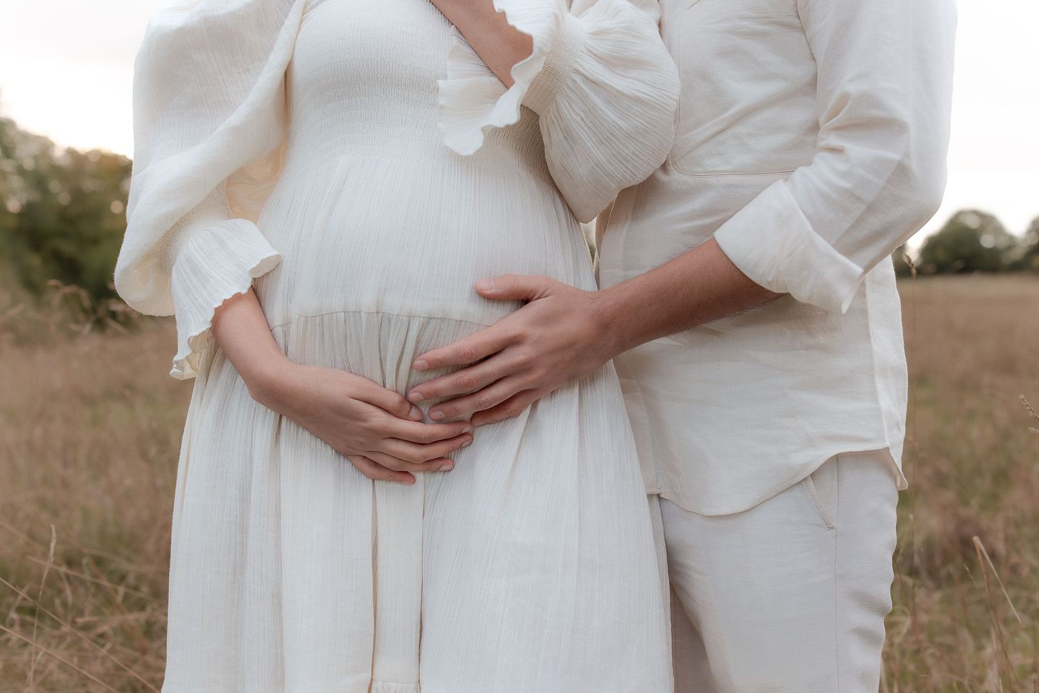 Couple in white outfits cradling pregnant belly in a serene field, capturing a tender maternity moment.