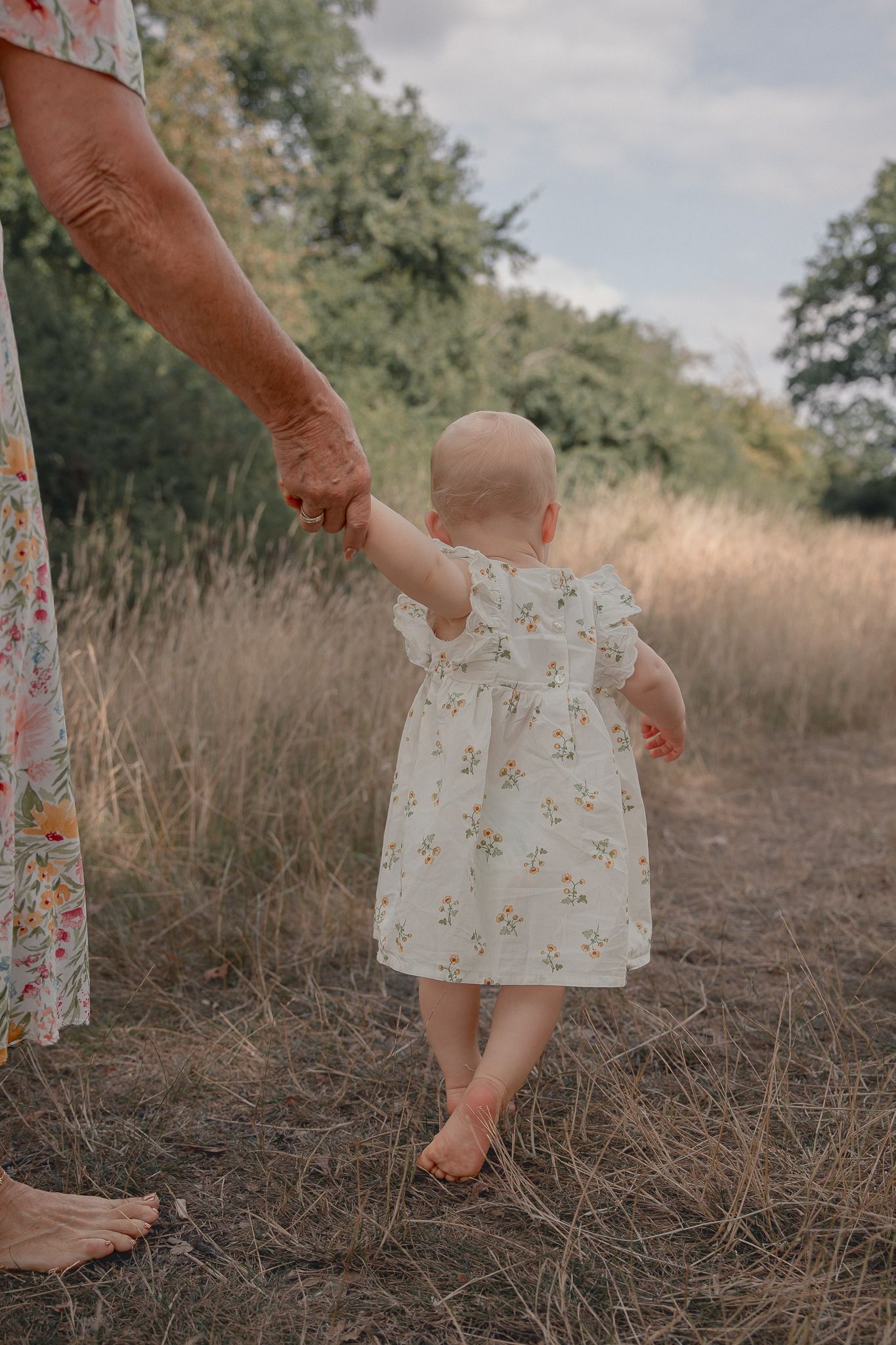 A baby in a floral dress takes first steps outdoors, holding an adult's hand in a golden field.