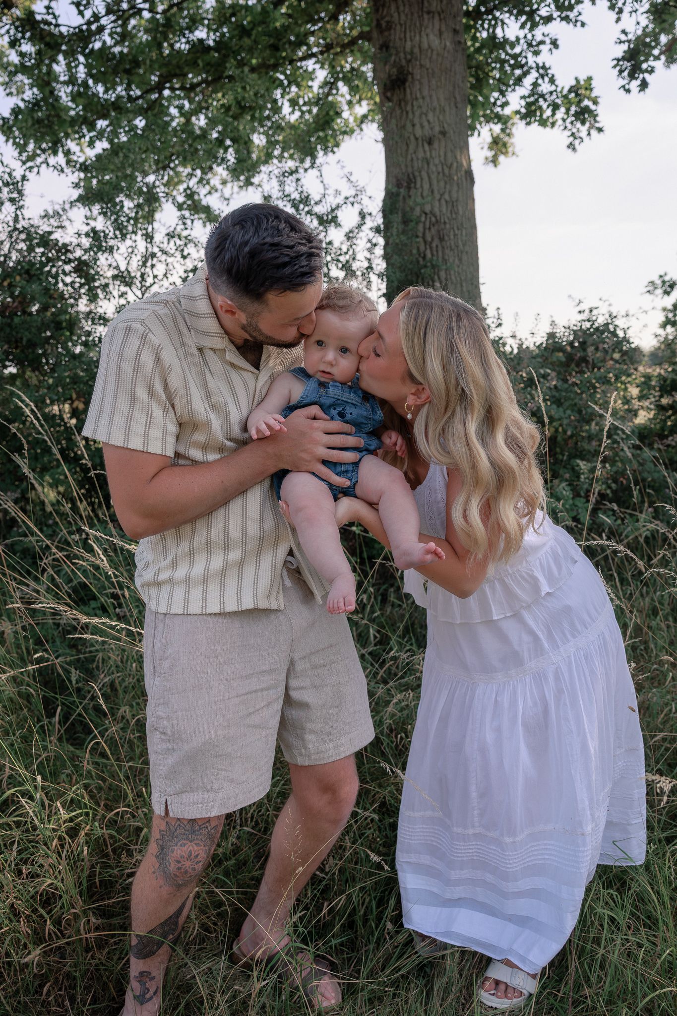 A couple kisses their baby outdoors near a tree in tall grass, mom in white dress, dad in beige shorts in Summertown, Oxford.