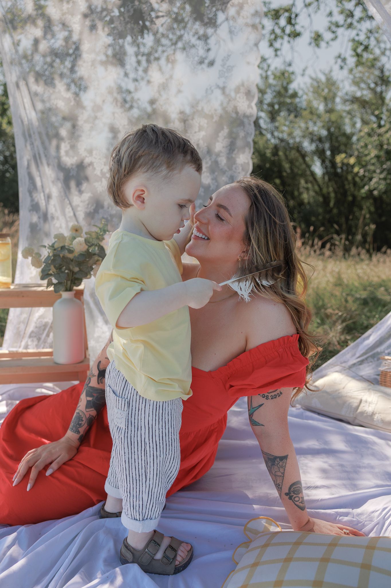 A smiling mother in a red dress shares a tender moment with her toddler son outdoors on a sunny day in Fairford, Cotswolds.