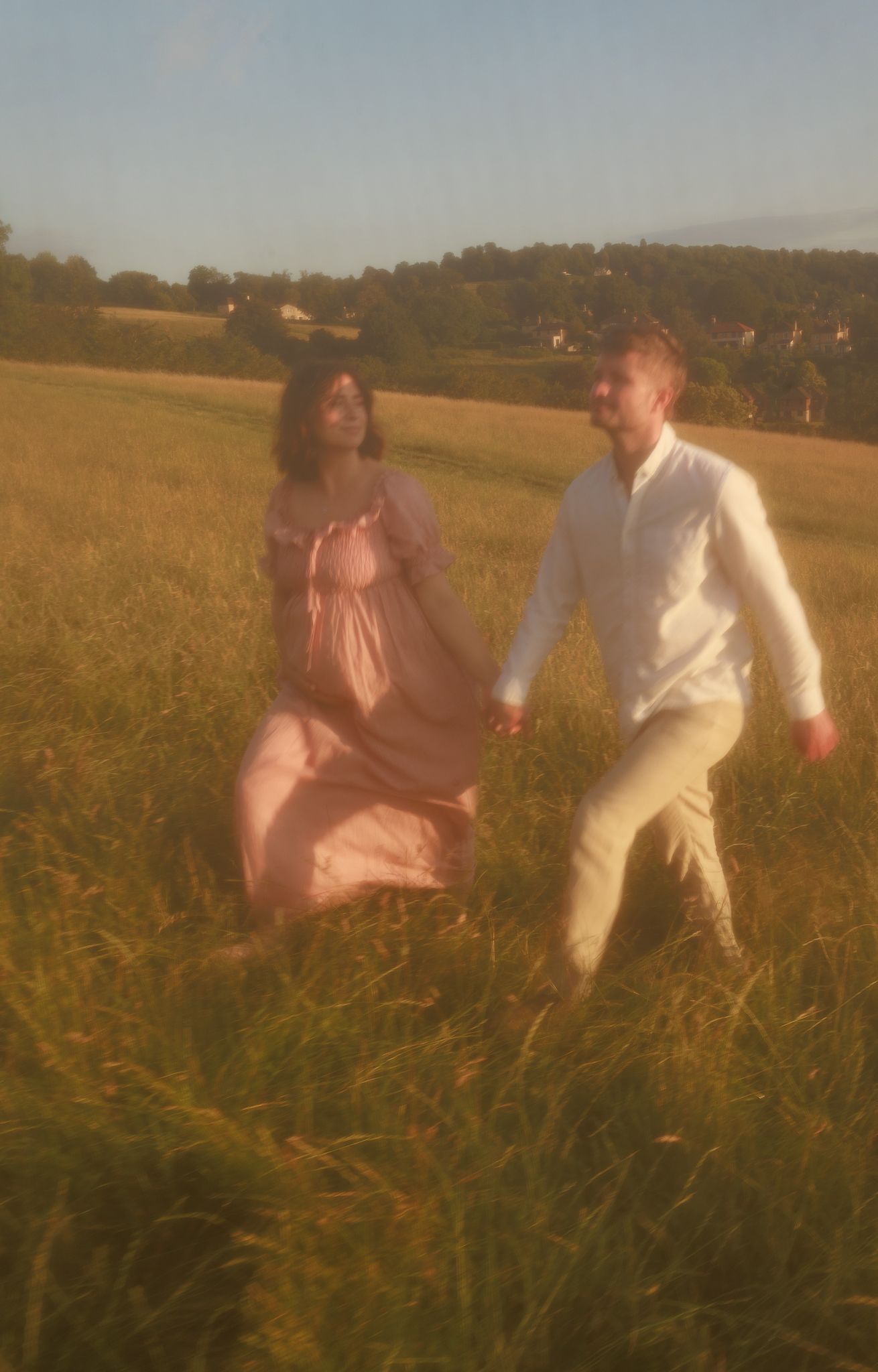 A couple holds hands while running through a golden sunlit field, the woman wearing a flowing pink dress for a Vintage Photoshoot in Oxford.