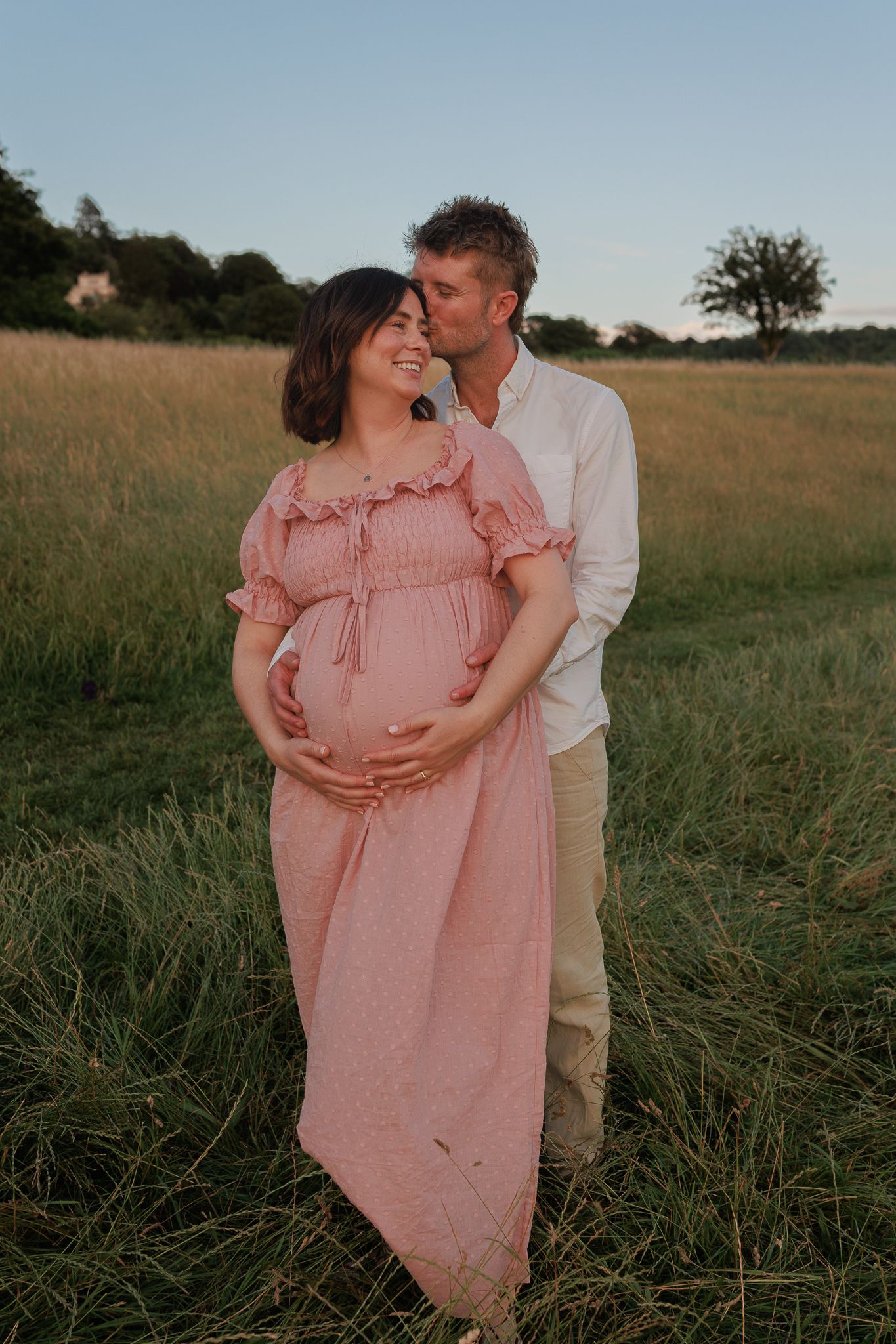 Man kisses pregnant woman in pink dress while she smiles in a sunlit grassy field during Maternity Photoshoot in Ascot.