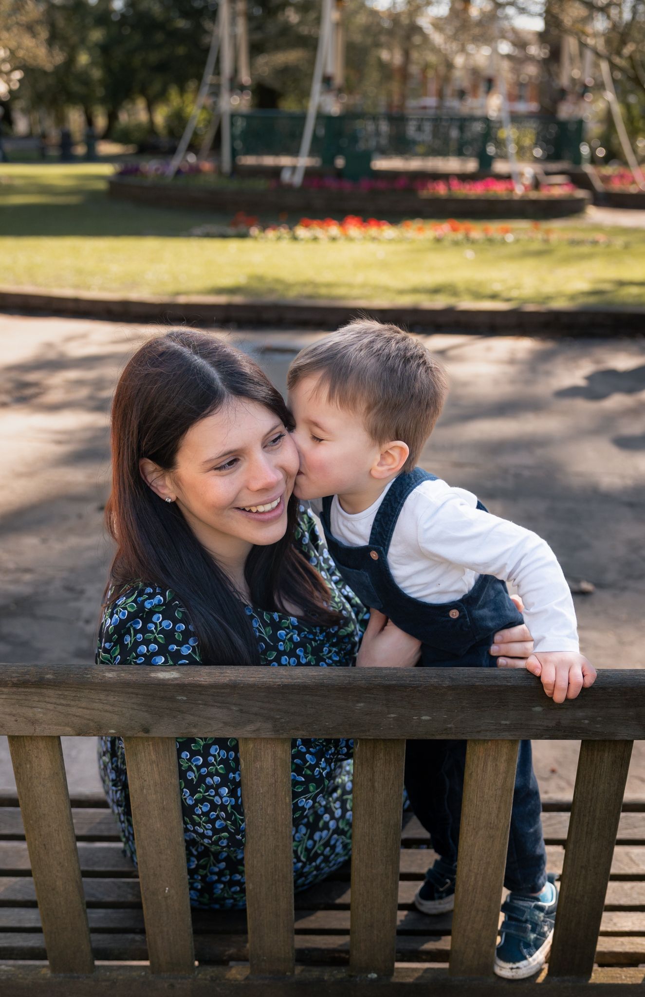 A young mother smiles as her toddler son kisses her cheek on a wooden park bench outdoors in North Cerney, Cotswolds.