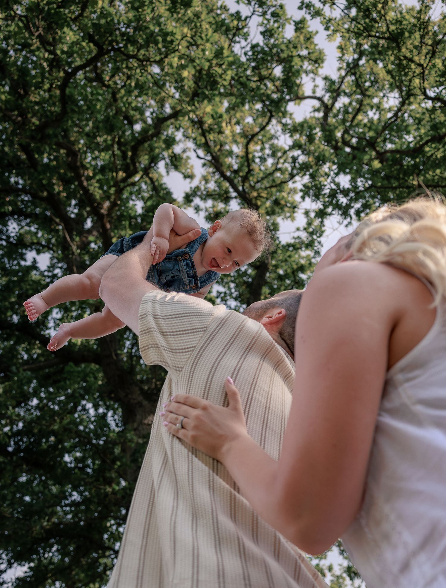 Parents joyfully lifting a laughing baby in the air outdoors, surrounded by lush green trees on a sunny day in Stow, Cotswolds.