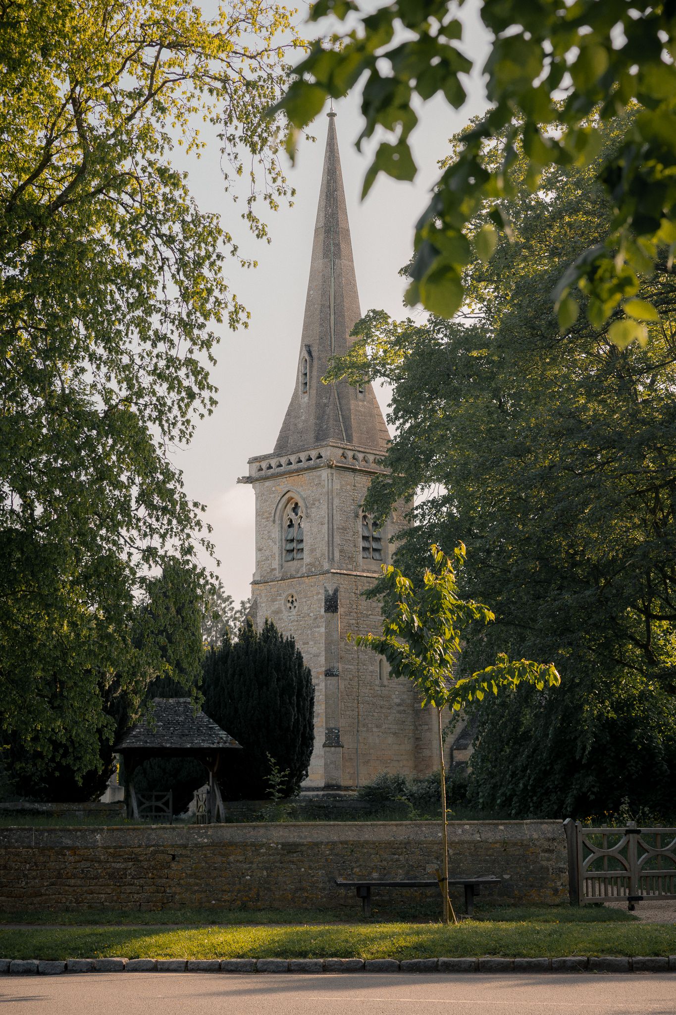 St Mary's church with tall pointed spire surrounded by lush green trees and a low stone wall in a peaceful English village.