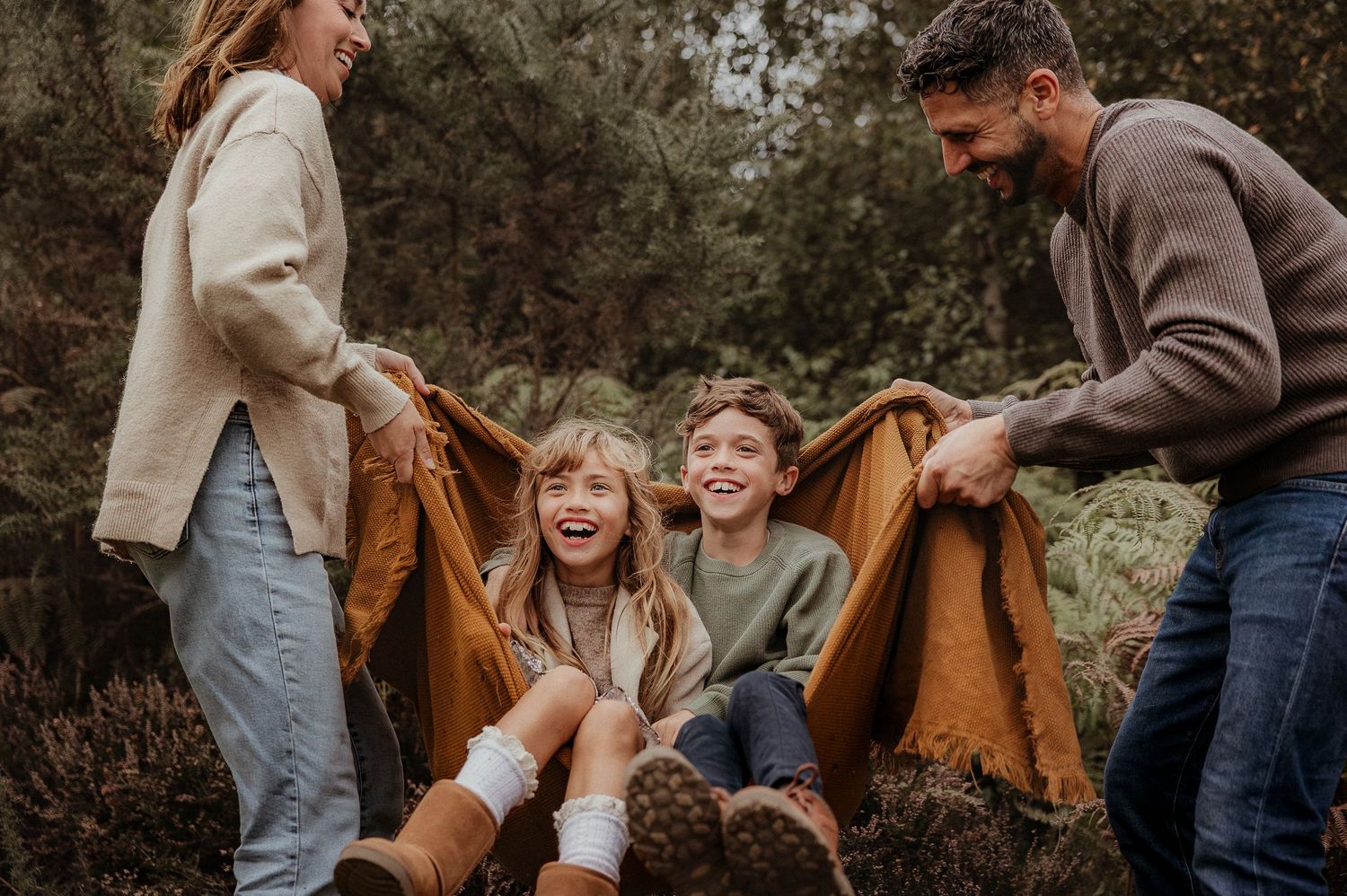 Family photoshoot of family laughing outdoors as parents swing two children in a brown blanket surrounded by lush greenery in Cirencester.