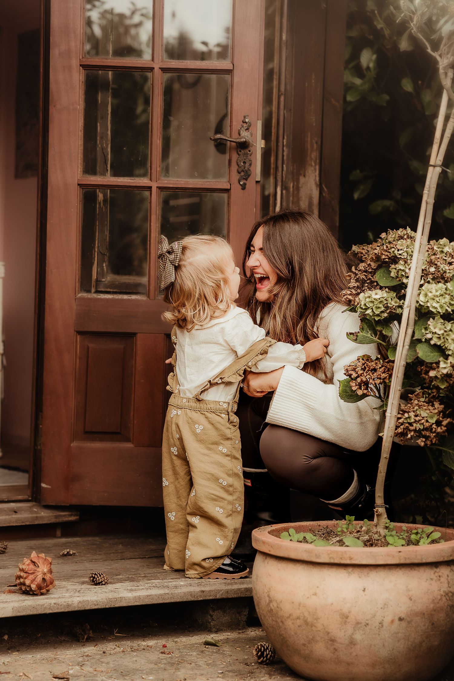 A joyful mother crouches by a wooden door, laughing with her toddler in tan overalls on a charming porch in Wroughton, Swindon.