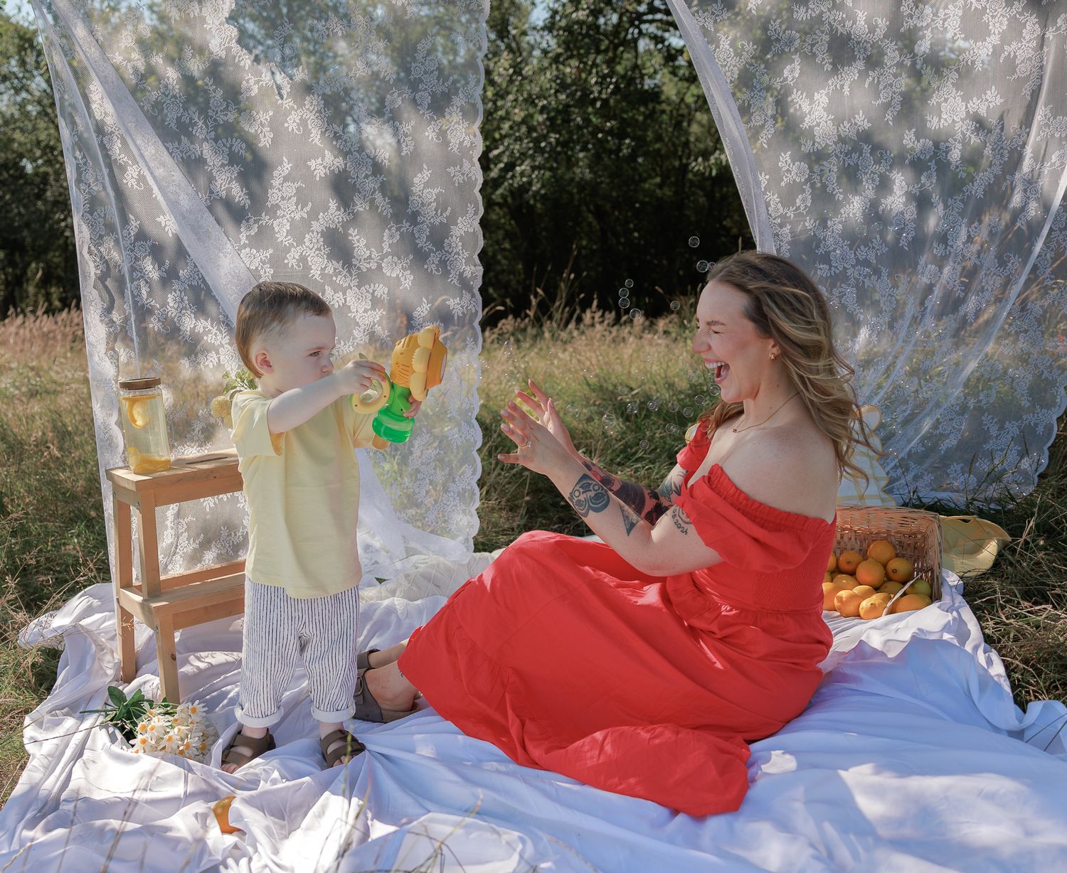 A laughing mother in a red dress plays with her toddler son at a sunny outdoor picnic in Fairford, Cotswolds.