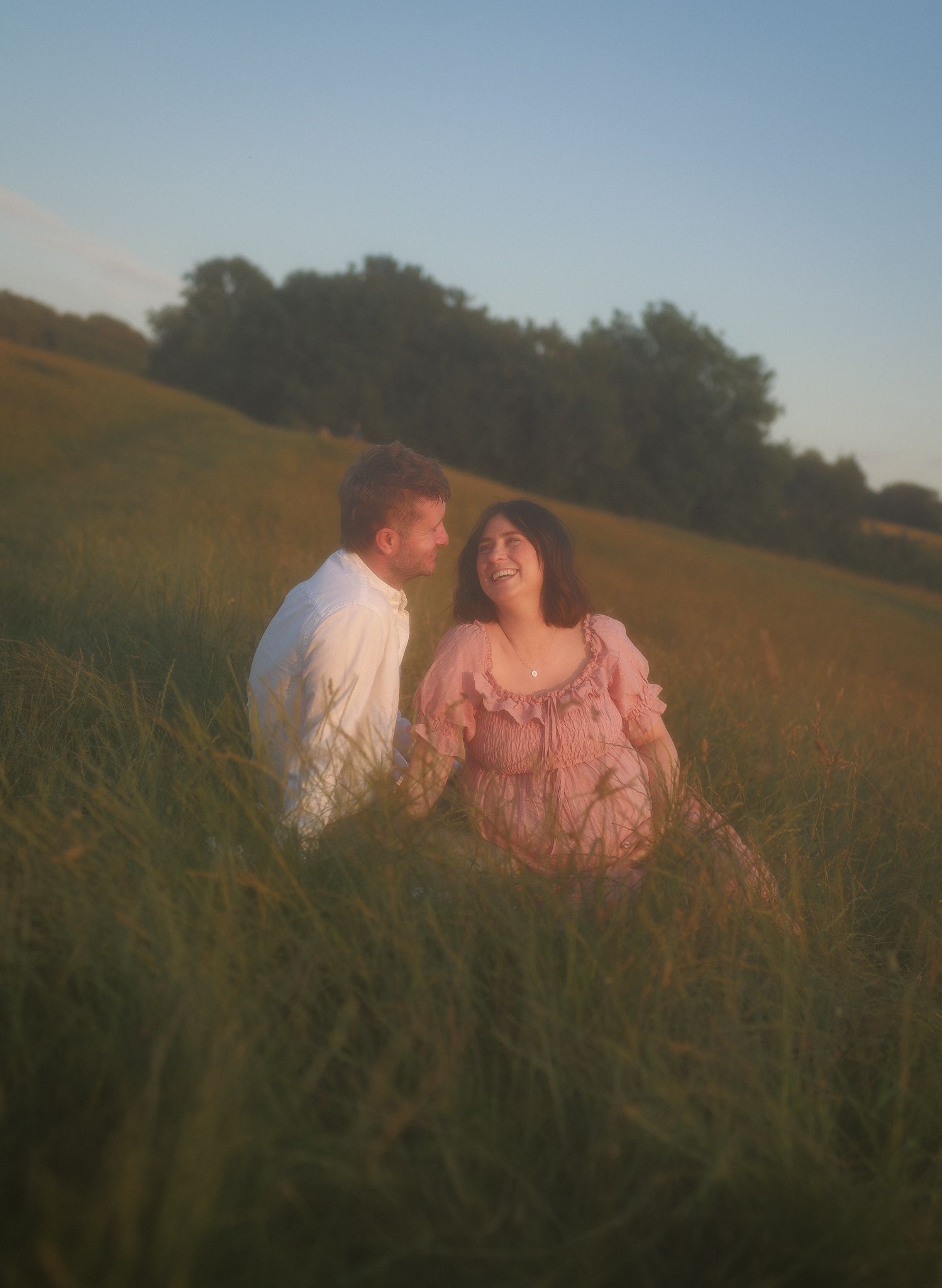 Couple sitting in tall grass, laughing together at golden hour looking over the Bath skyline. Maternity Photoshoot Bath.