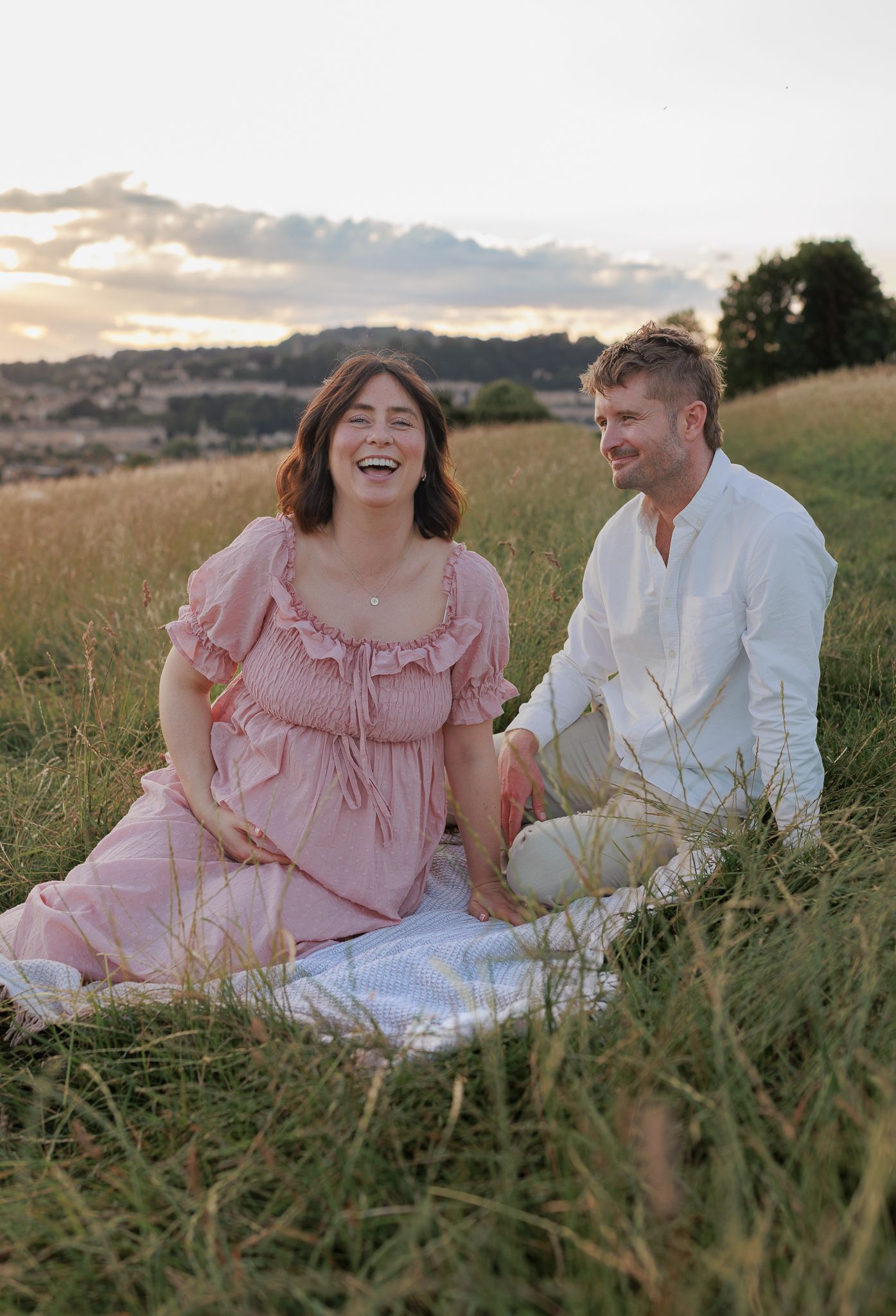 A laughing pregnant woman sits with a man in white shirt in a golden field at sunset for a Maternity Photoshoot in Cotswolds, UK.