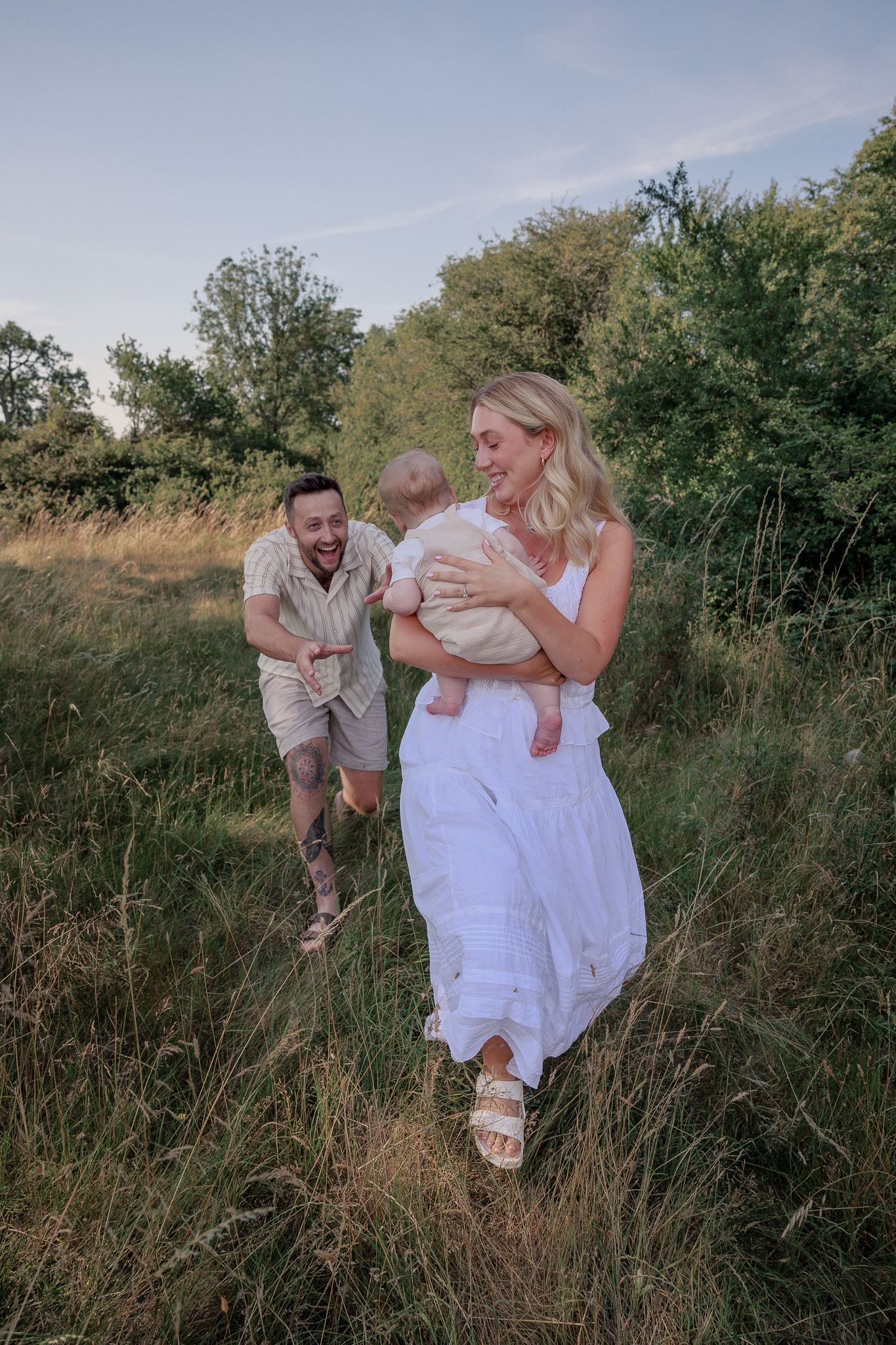 Happy young family with baby walking in green meadow, mother in white dress, father laughing behind her in North Cerney, Cotswolds.