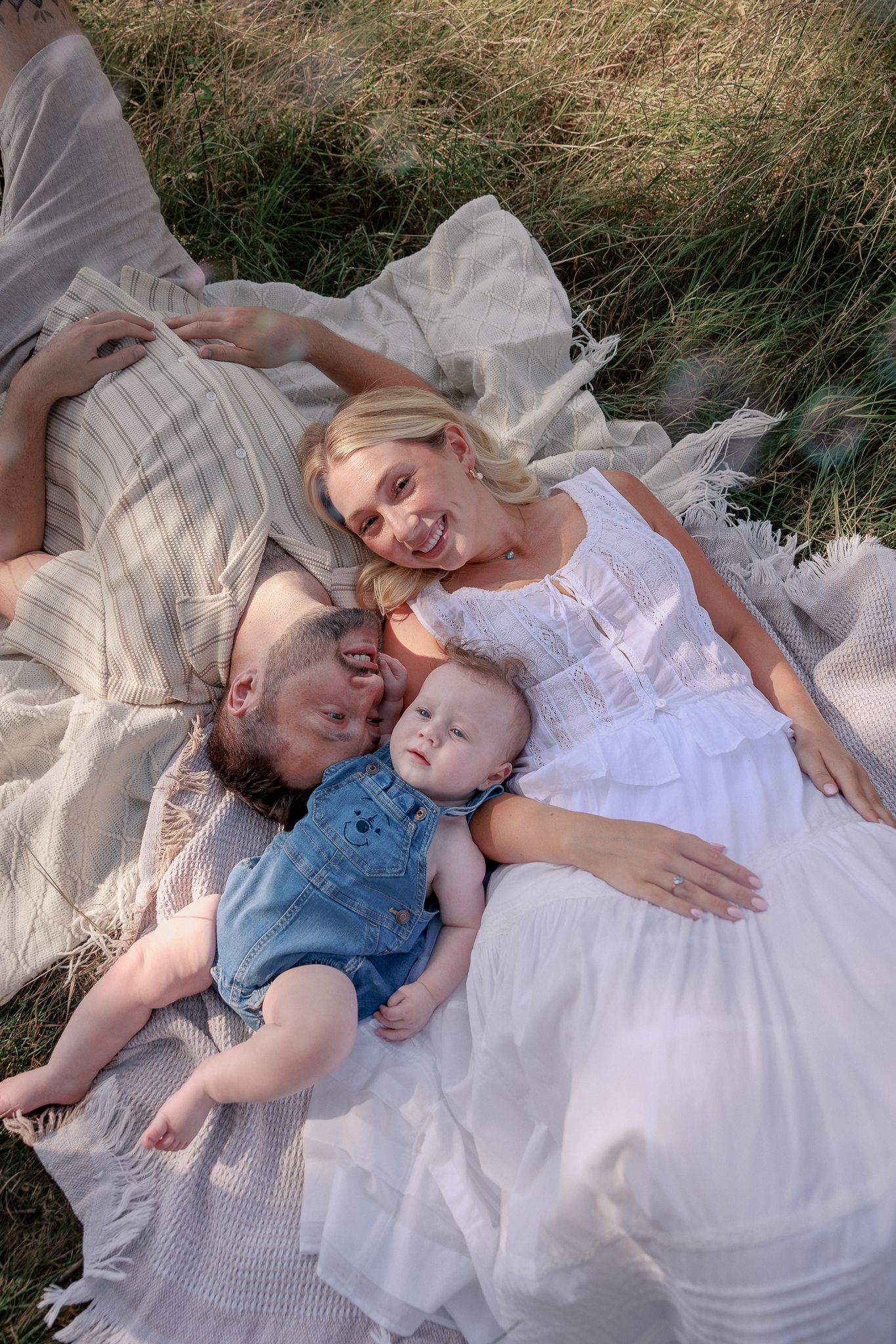 Family of three lying on a blanket in a grassy field, parents smiling at baby in denim overalls in Stow, Cotswolds.