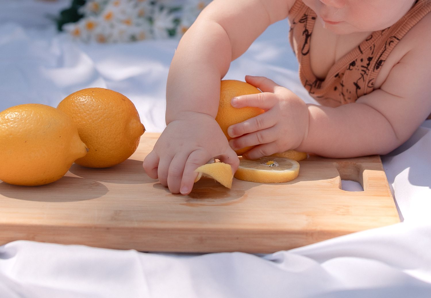 Baby reaching for fresh lemons on a wooden cutting board, with sliced citrus fruit on a white blanket in Burford, Cotswolds.