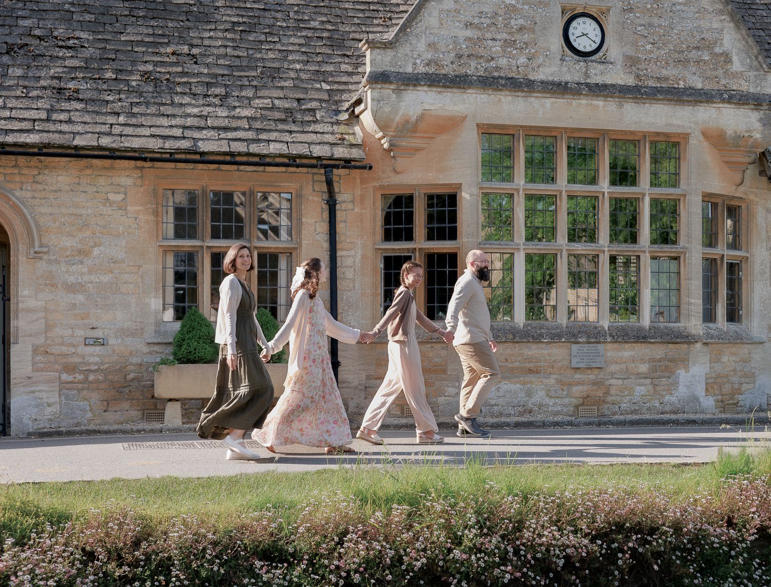Family of four walks hand-in-hand past a historic stone manor house on a sunny day.
