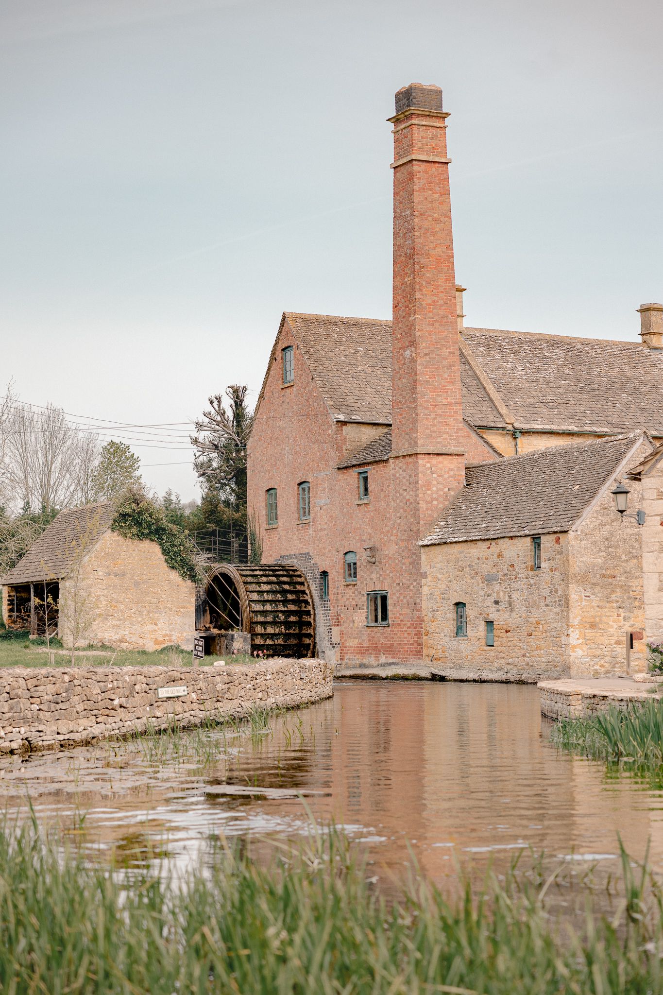 Lower Slaughter Museum a historic brick watermill with chimney stack beside a calm river, reflecting in water.