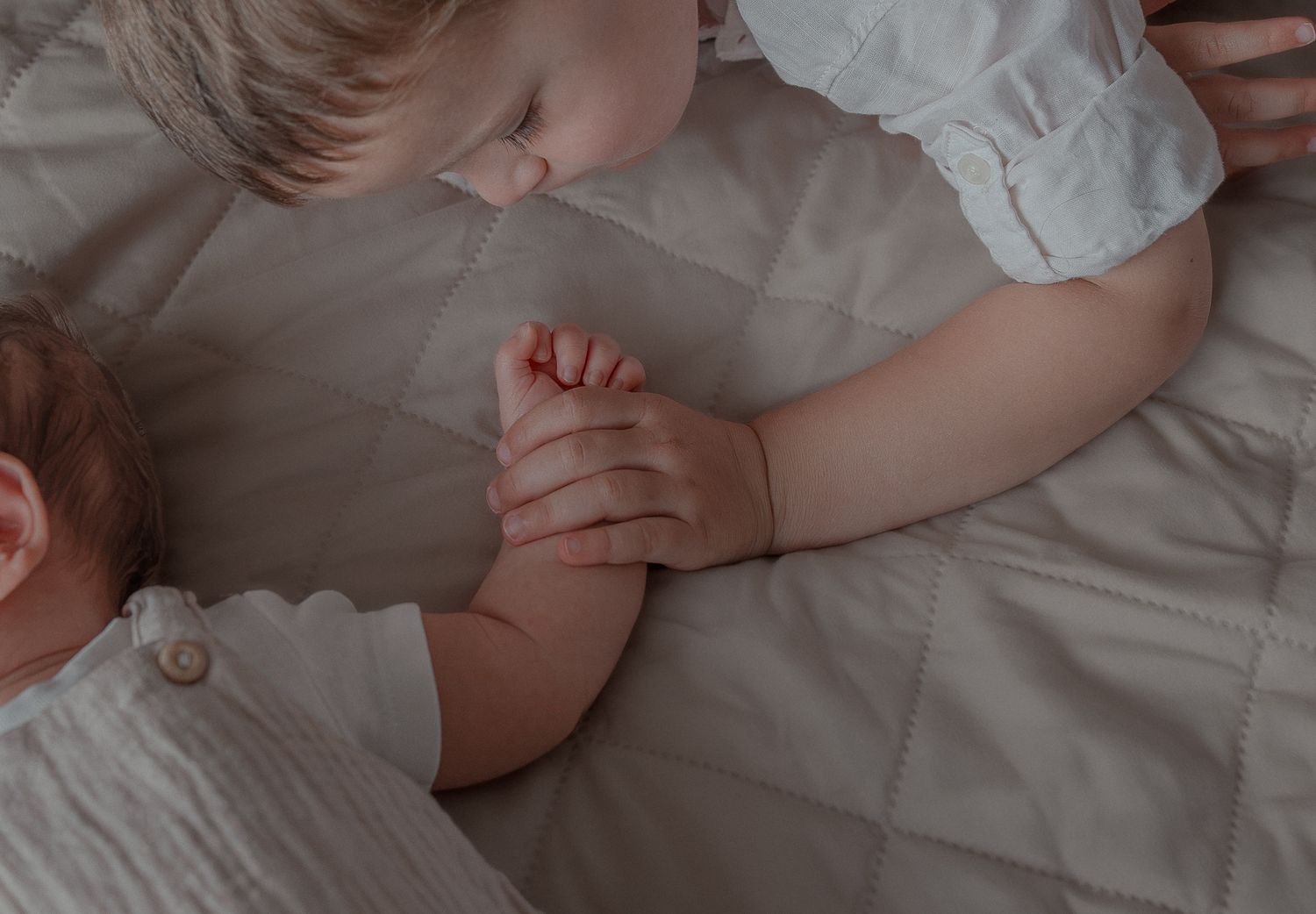 Toddler in white shirt holds newborn baby's hand on a quilted white blanket, siblings bonding moment in Clifton, Bristol.