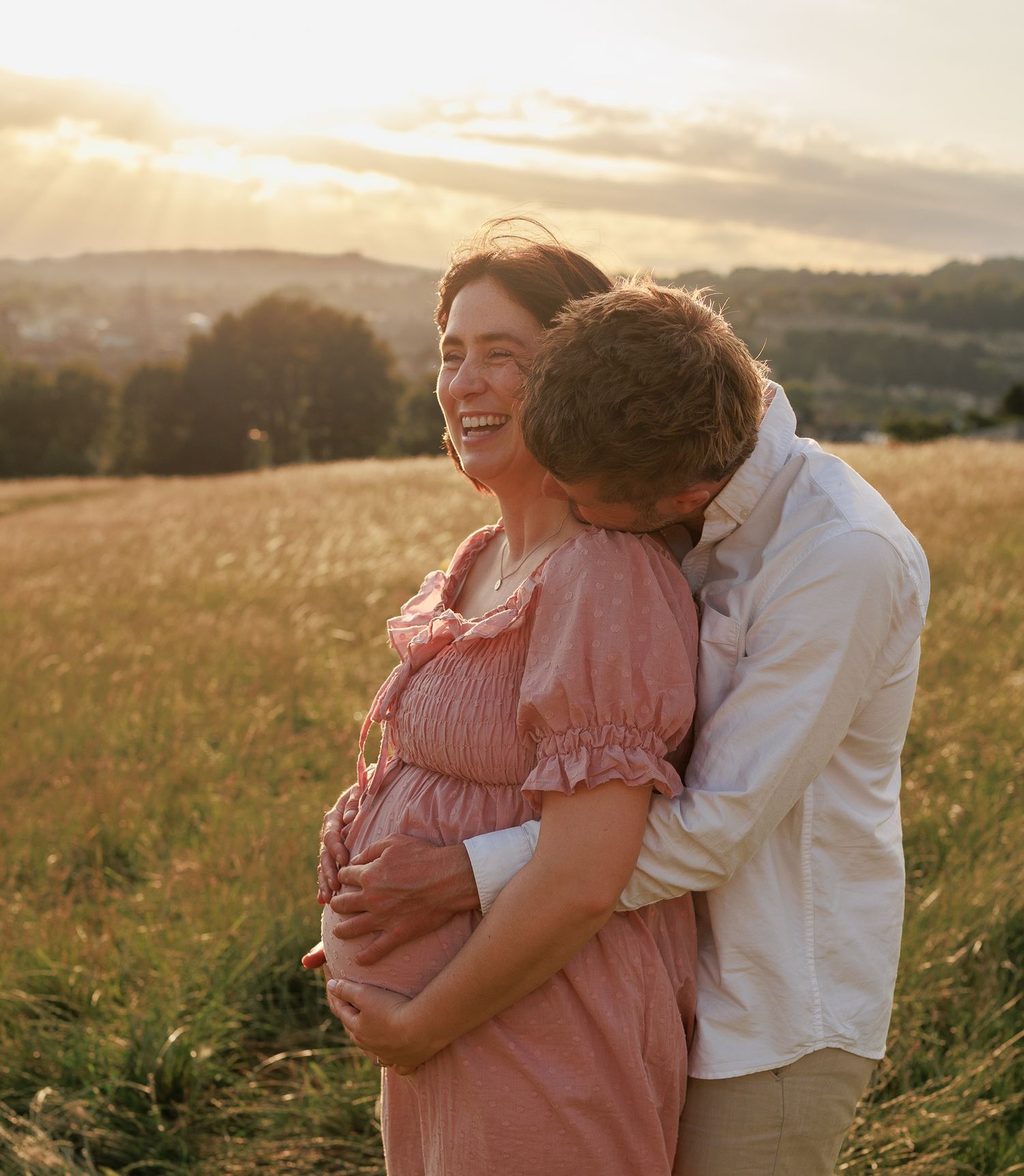 Pregnant woman in pink dress laughs as partner embraces her in golden sunset field for a Maternity Photoshoot in Wiltshire, UK.