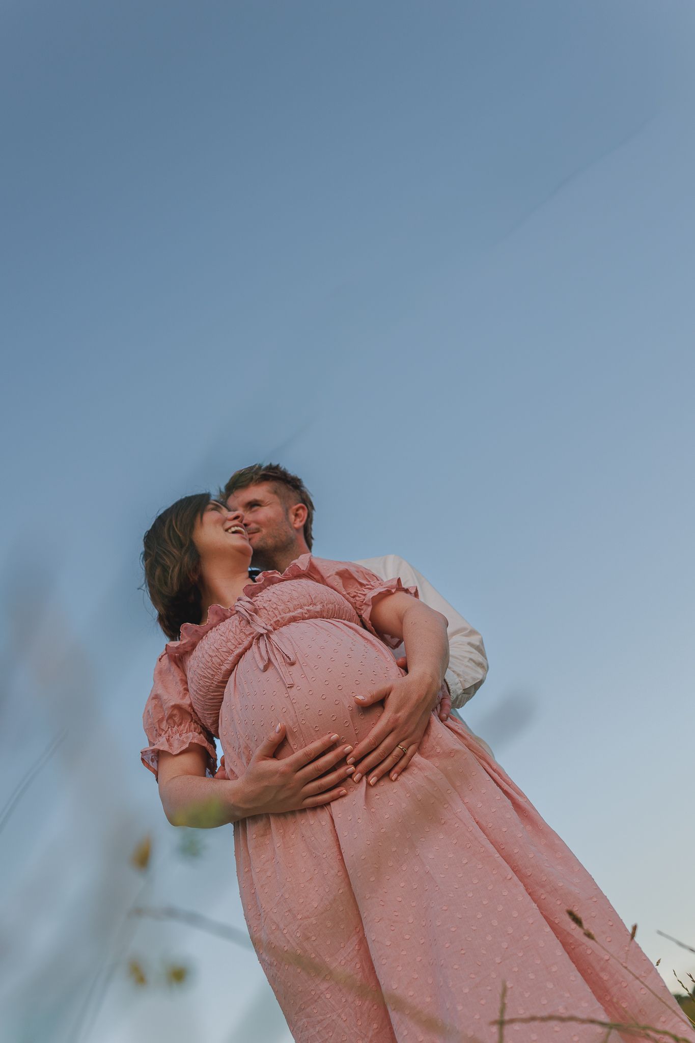 A couple embraces outdoors, the pregnant woman in a pink dress smiling as her partner holds her baby bump against a blue sky in Datchet.