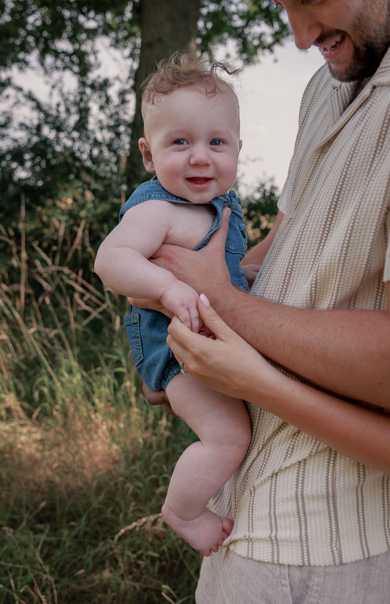 Smiling baby in denim overalls being held by a man outdoors in a green field in Stow, Cotswolds.