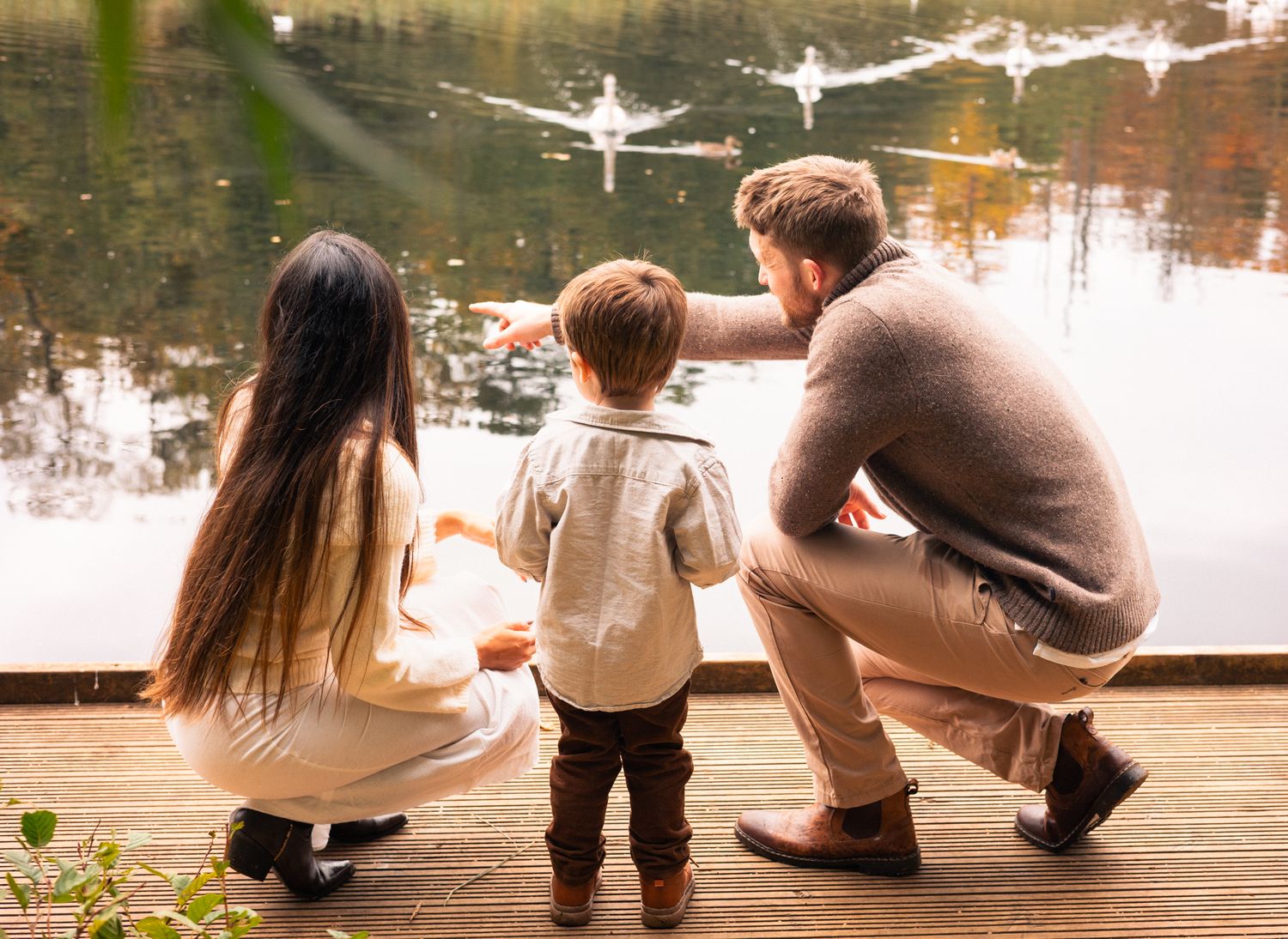 Family sitting together on a dock watching swans swim in a lake during Autumn in Cotswolds and Wiltshire, Chippenham.