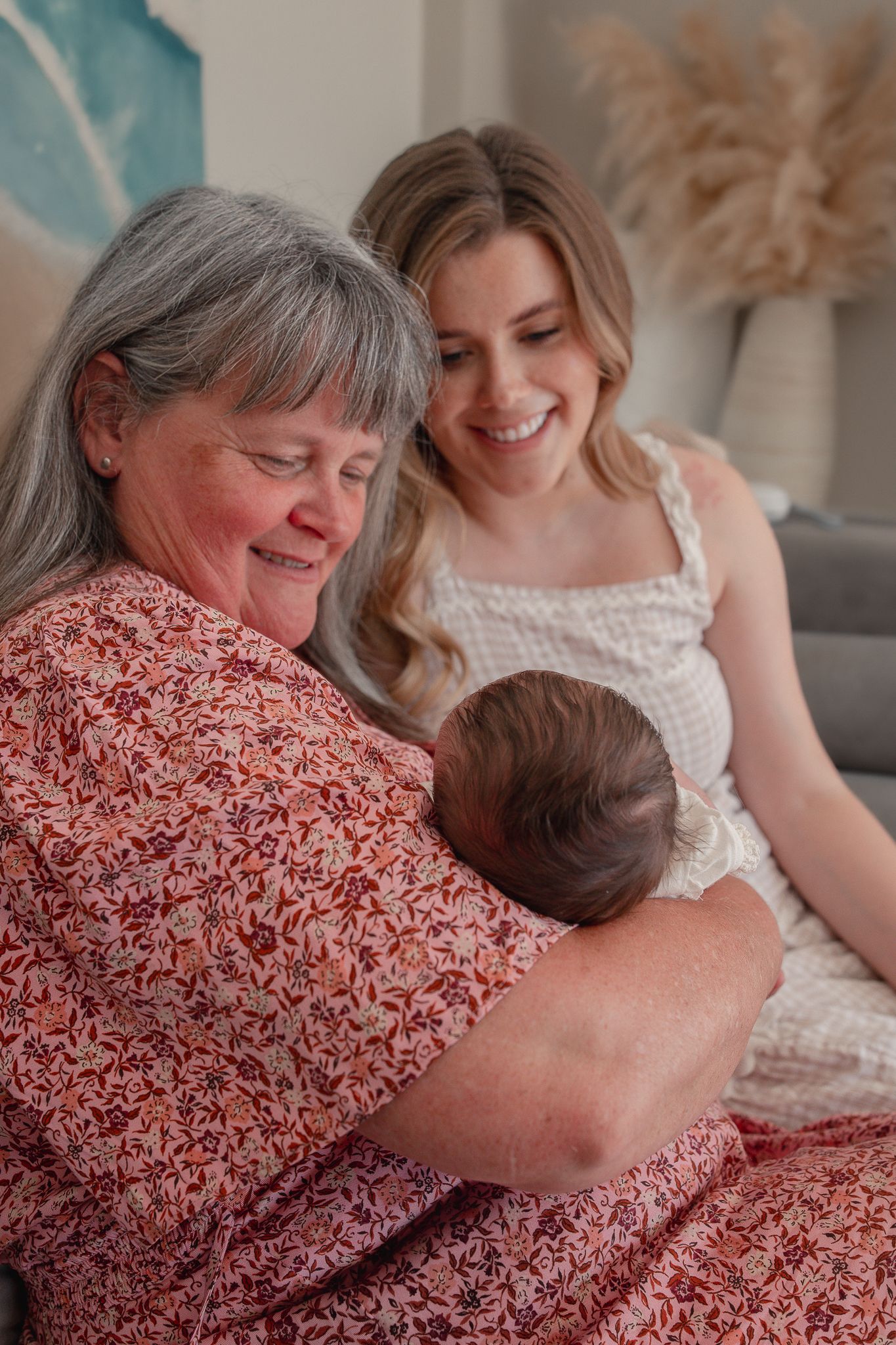 Grandmother holding newborn baby while young mother smiles warmly beside her on a cozy living room sofa for a Newborn Photoshoot, Bath, UK.