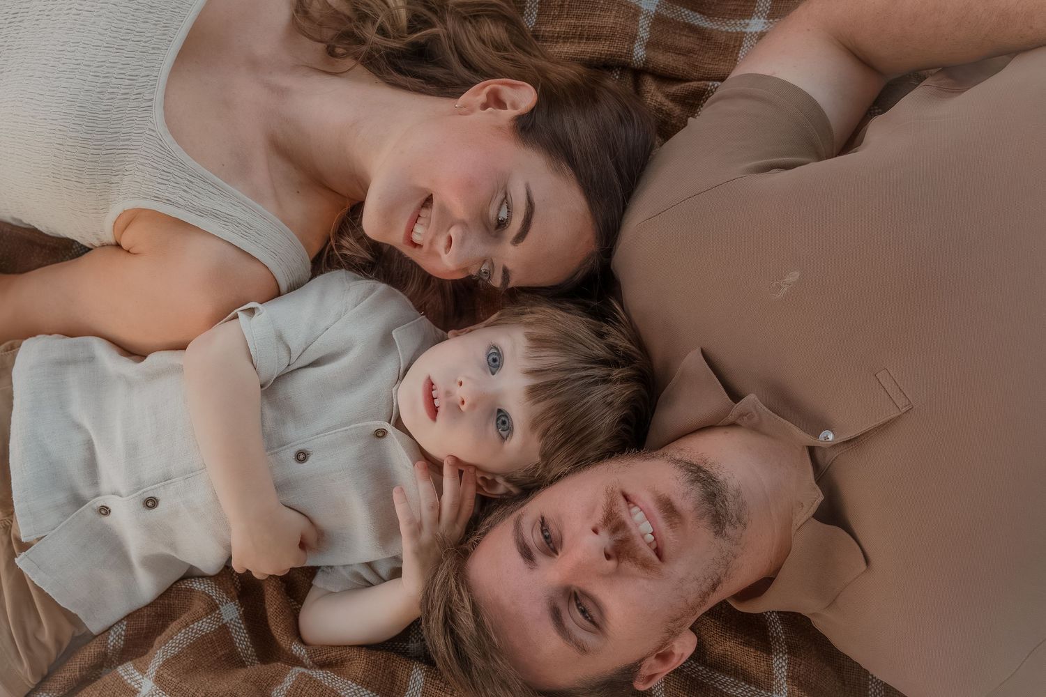 A happy young family lies together on a blanket, smiling up at the camera with their baby between them in Bath, Somerset..