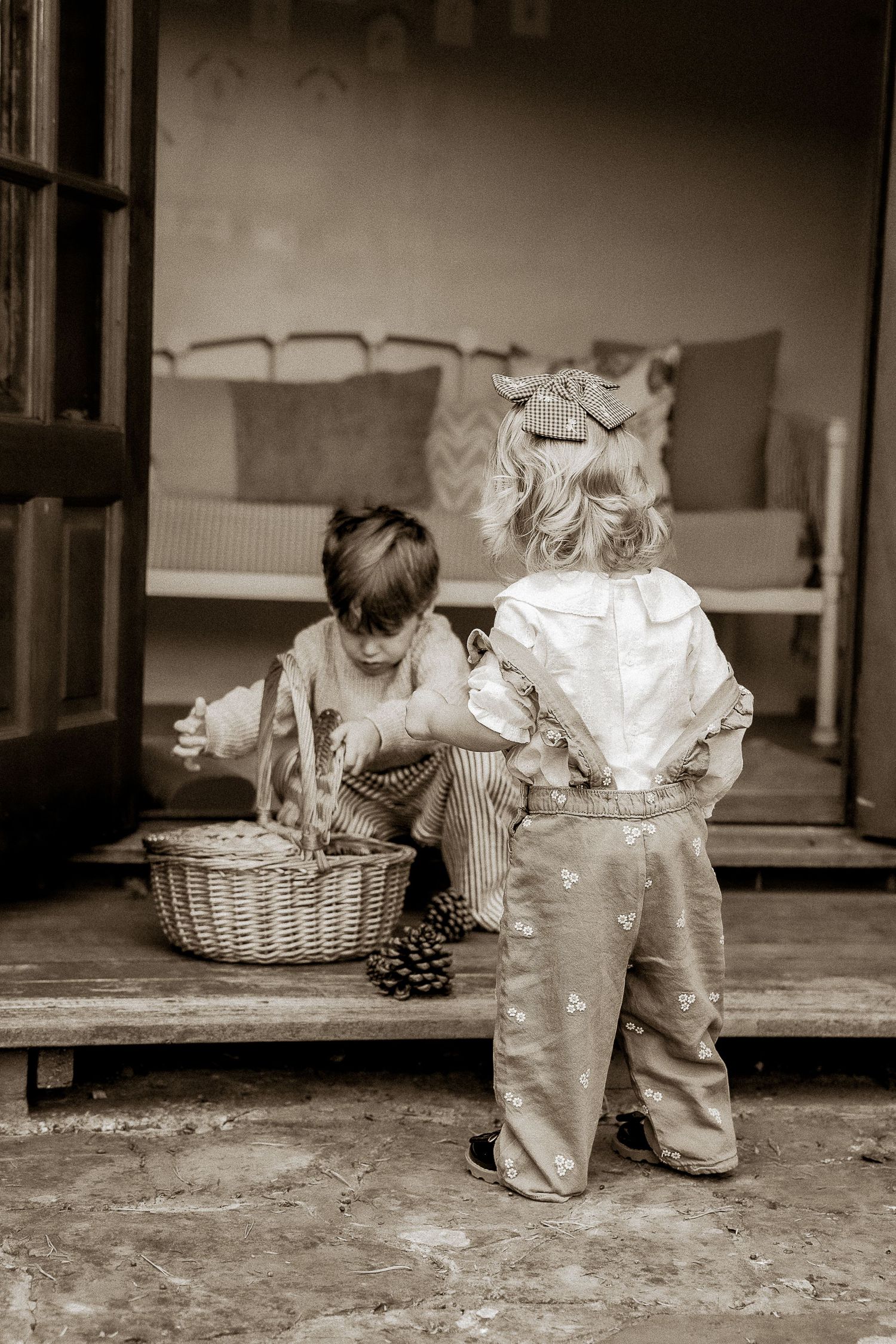 Two young children in vintage clothing examine a wicker basket on porch steps in sepia-toned photo in Lacock.
