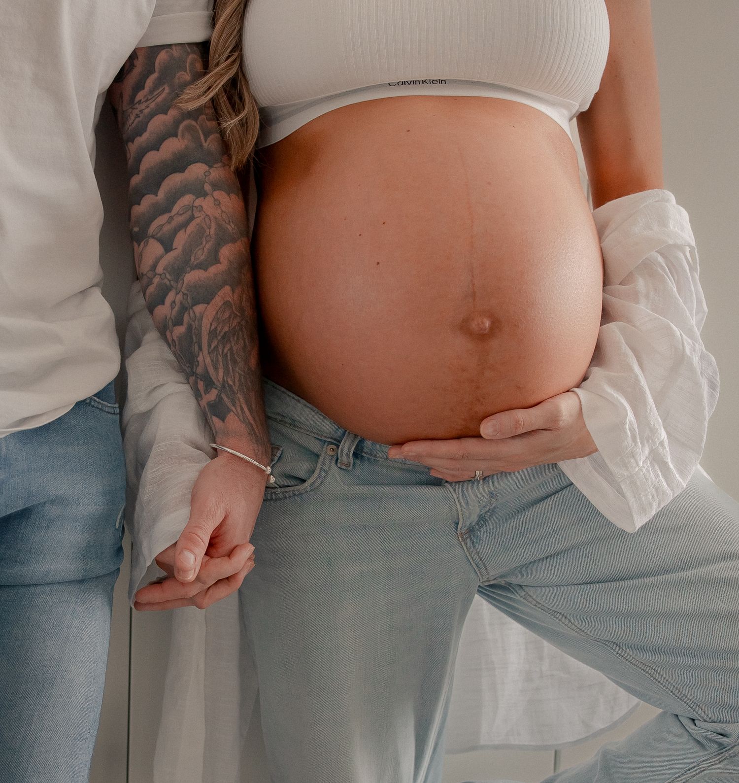 Tattooed partner embracing pregnant woman's bare baby bump, both wearing casual jeans and white tops in maternity portrait in Fairford.