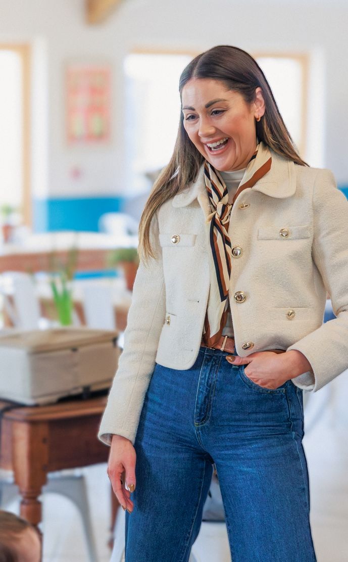 Woman in cream jacket and blue jeans smiling and laughing at a Mum and baby event in Cirencester. Cotswolds, Roots and Seeds.