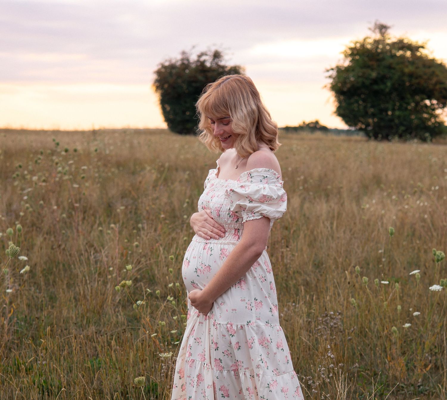 A mum to be in a floral dress stands in a golden field at sunset cradling baby bump in Asthall.