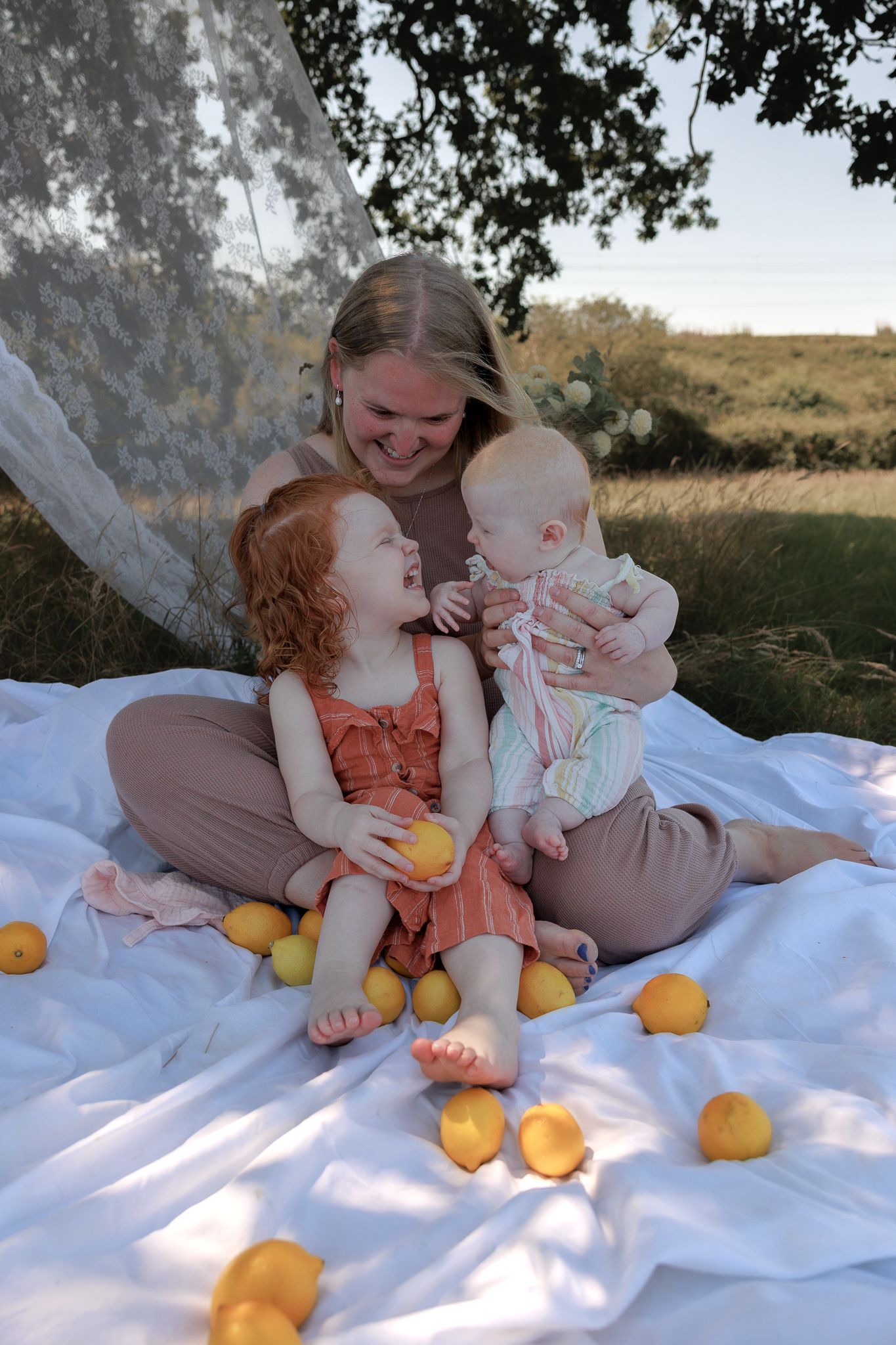 A smiling mother sits on a white blanket outdoors with her two young children surrounded by fresh lemons in Henley-on-Thames, Oxfordshire.
