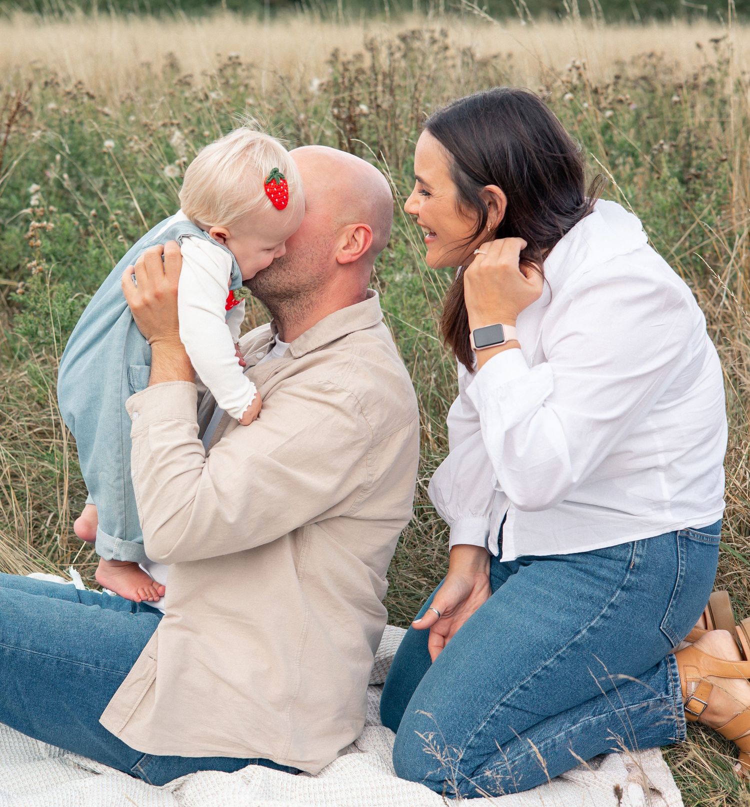 Family portrait of parents with baby sitting outdoors in a field during golden hour.