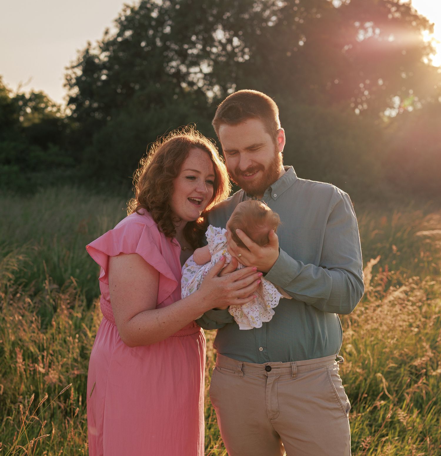 A couple stands in a sunlit field, smiling at their newborn baby, with golden hour light glowing behind them in Reading.
