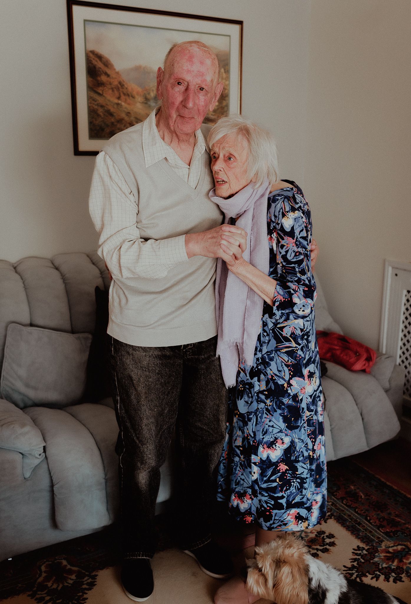 Elderly couple embracing warmly in a cozy living room with a framed landscape painting on the wall behind them in Broadway.