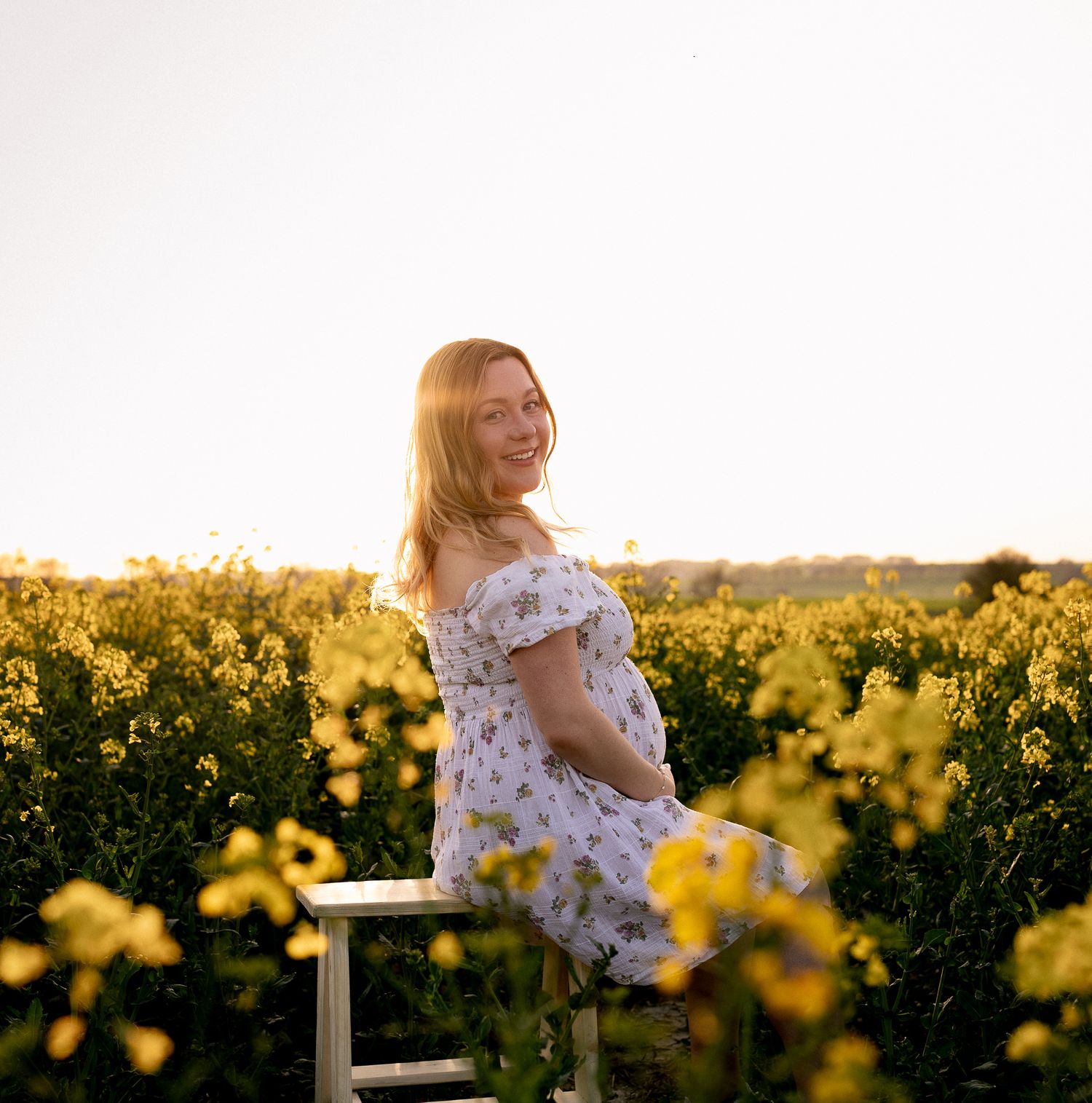 Pregnant woman in floral dress sitting in yellow rapeseed field, smiling in golden sunset light in Henley-on-Thames.