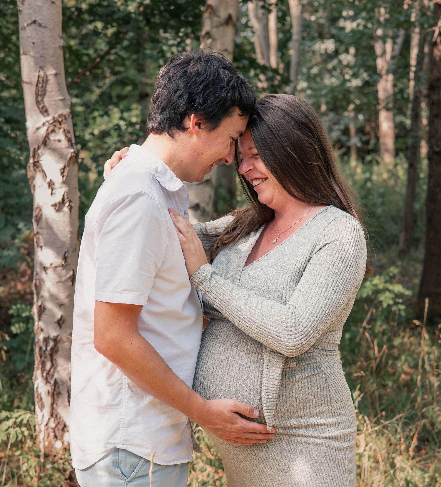 Couple sharing a tender moment outdoors, woman in grey maternity dress, surrounded by lush green trees in Worcestershire.