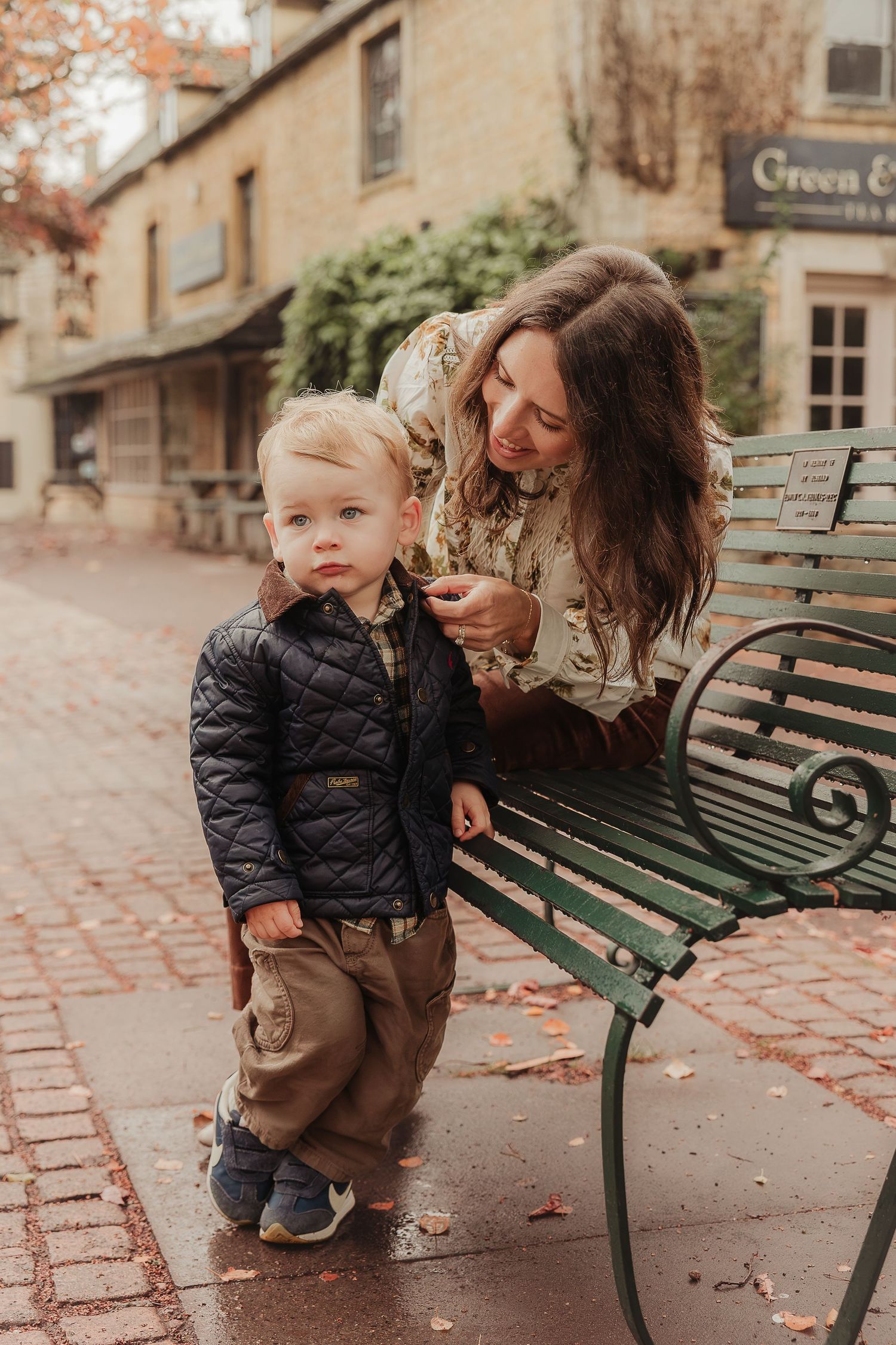 Smiling woman with long brown hair adjusts the jacket of a toddler in a quilted coat beside a green bench on a brick-paved street in autumn.