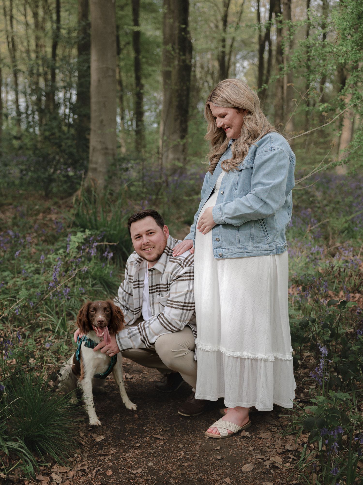 Couple in woodland maternity photoshoot with spaniel dog, woman in white dress and denim jacket cradling baby bump.