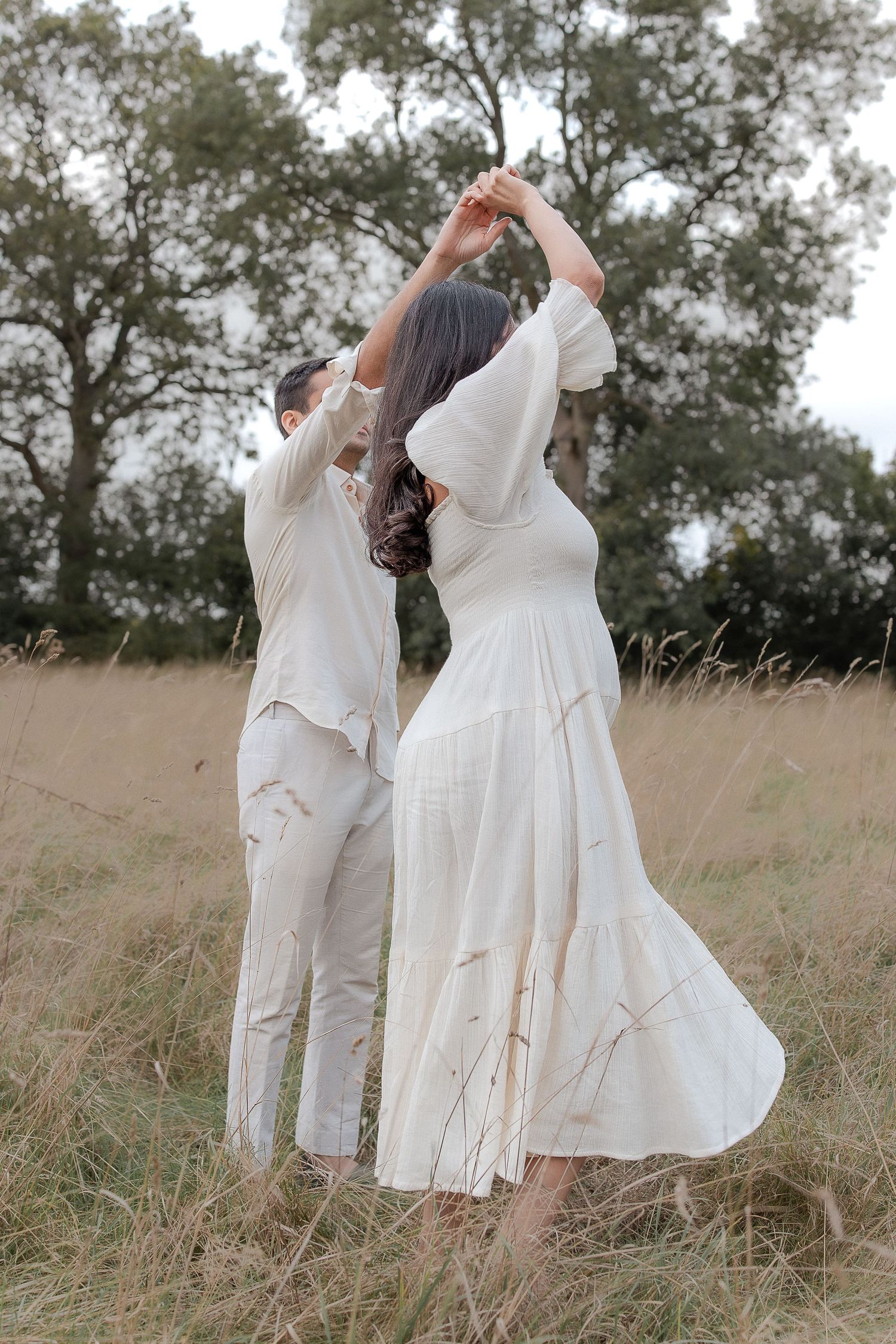 A couple in white outfits dancing together in a golden meadow with trees in the background.