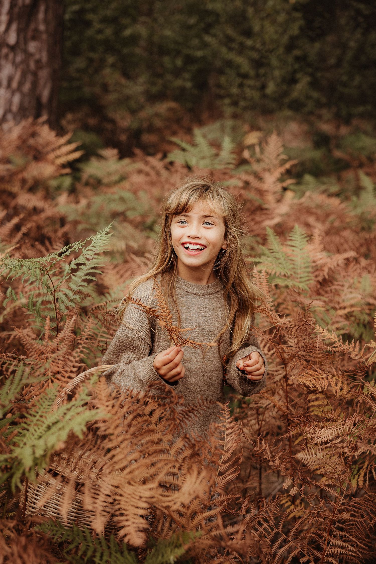Family Photoshoot of smiling girl with long blonde hair standing among autumn ferns in a forest in Moreton-On-Marsh.