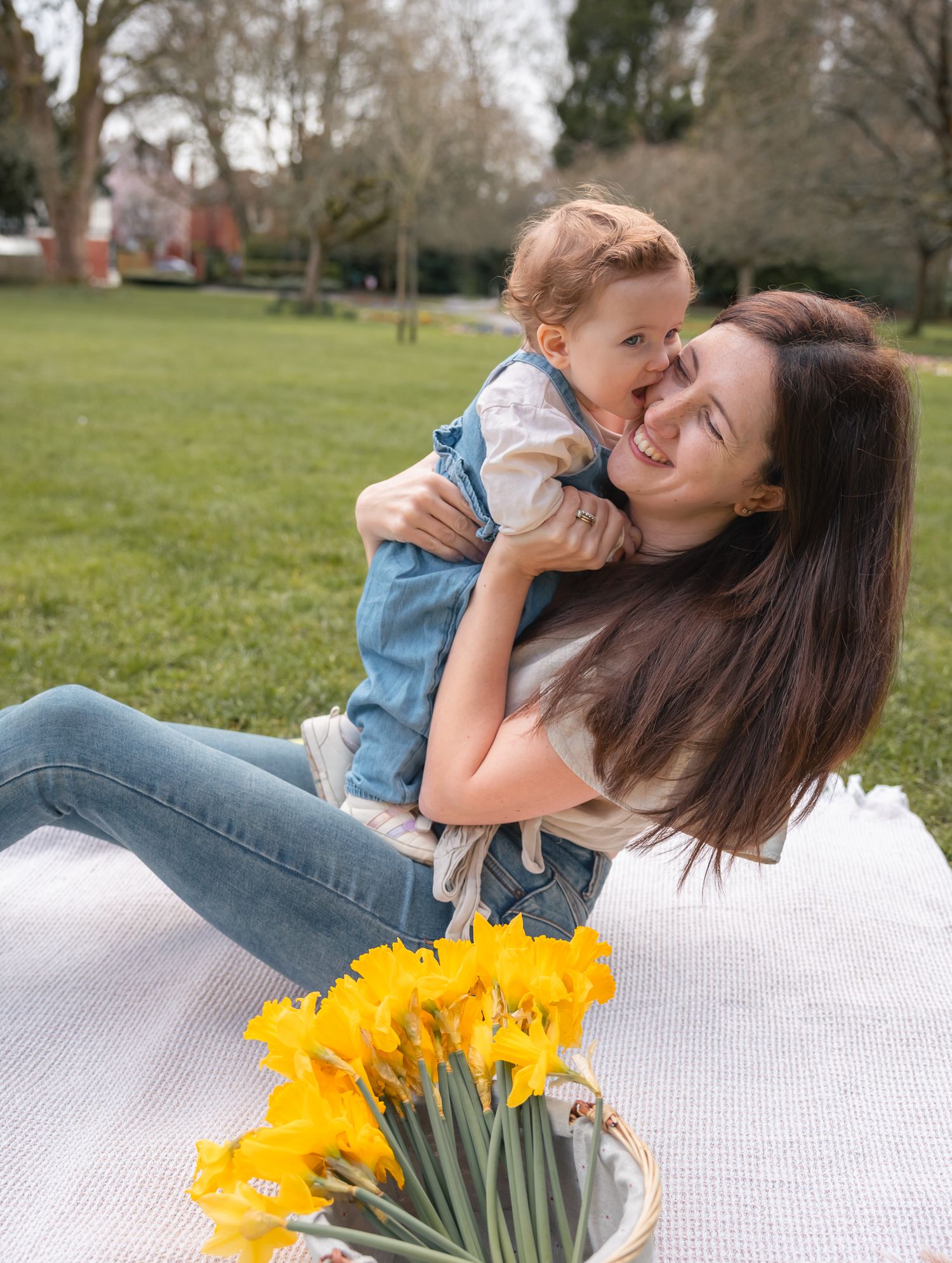 A smiling mother holds her baby on a sunny spring picnic blanket with yellow daffodils in a lush green park in South Cerney, Cotswolds.
