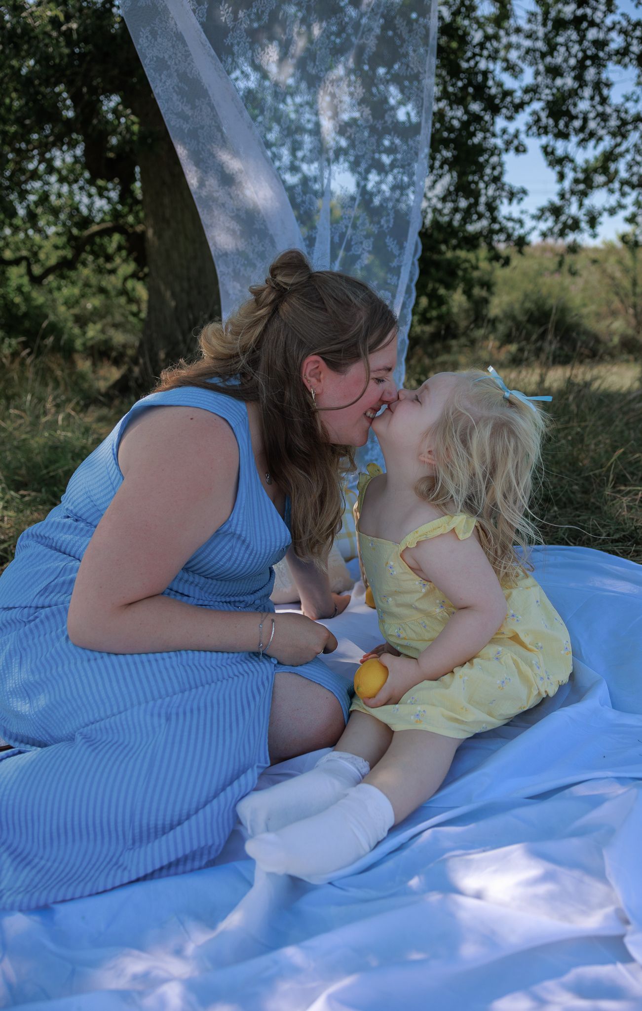 A mother in a blue dress shares a sweet kiss with her toddler daughter in a yellow dress during an outdoor picnic in Bibury, Cotswolds.