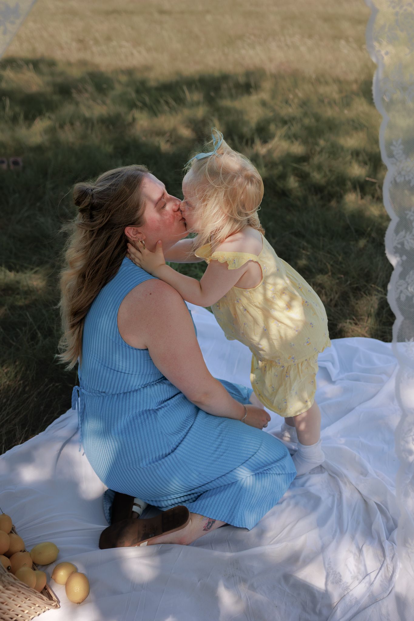 A mother in a blue dress kisses her toddler in a yellow outfit on a white blanket outdoors in Bibury, Cotswolds.