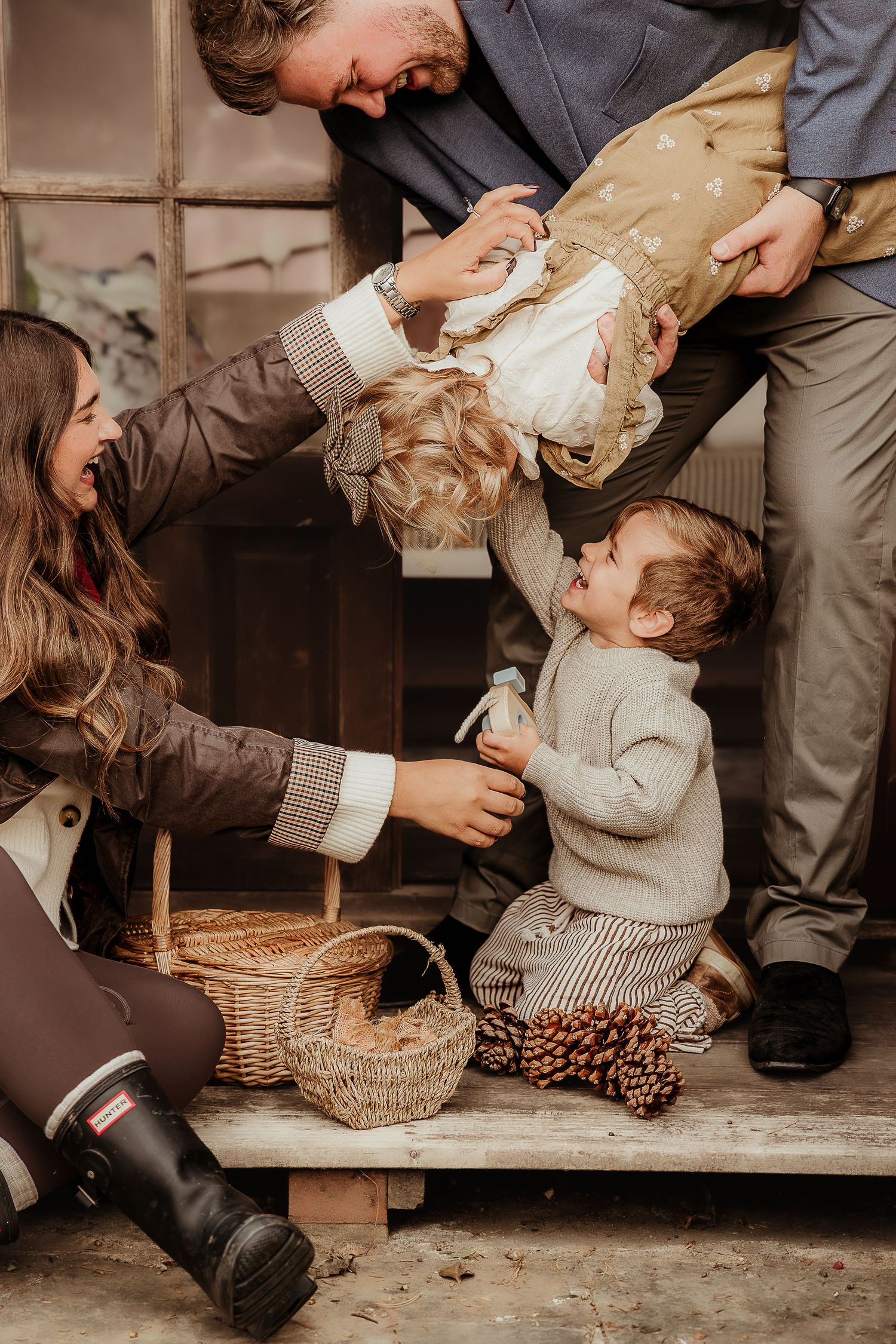 Family Photoshoot with two young children playing on rustic wooden porch steps with pine cones in Autumn in Bourton-On-Water.