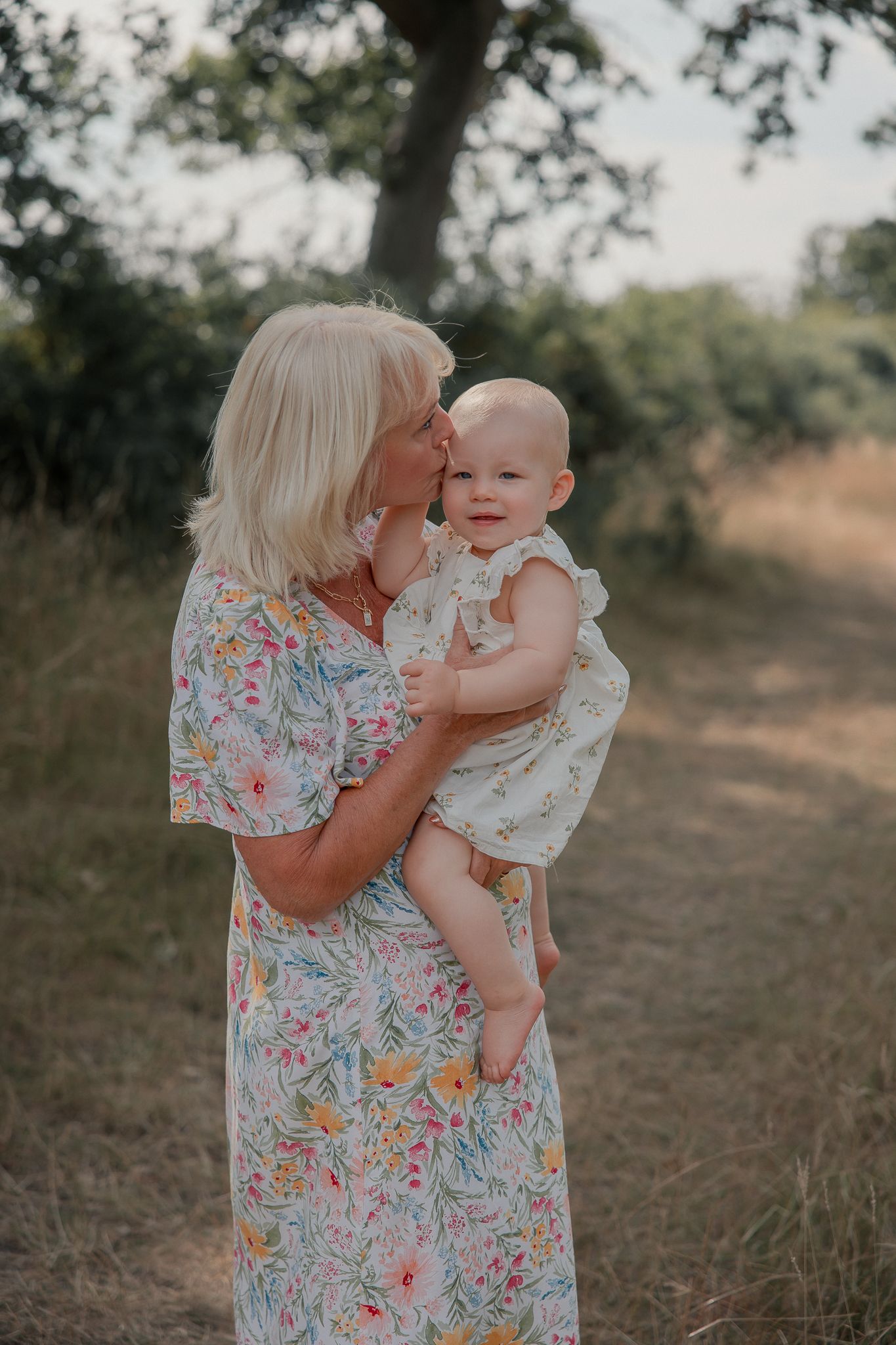 Blonde woman in floral dress kissing baby girl outdoors in nature.