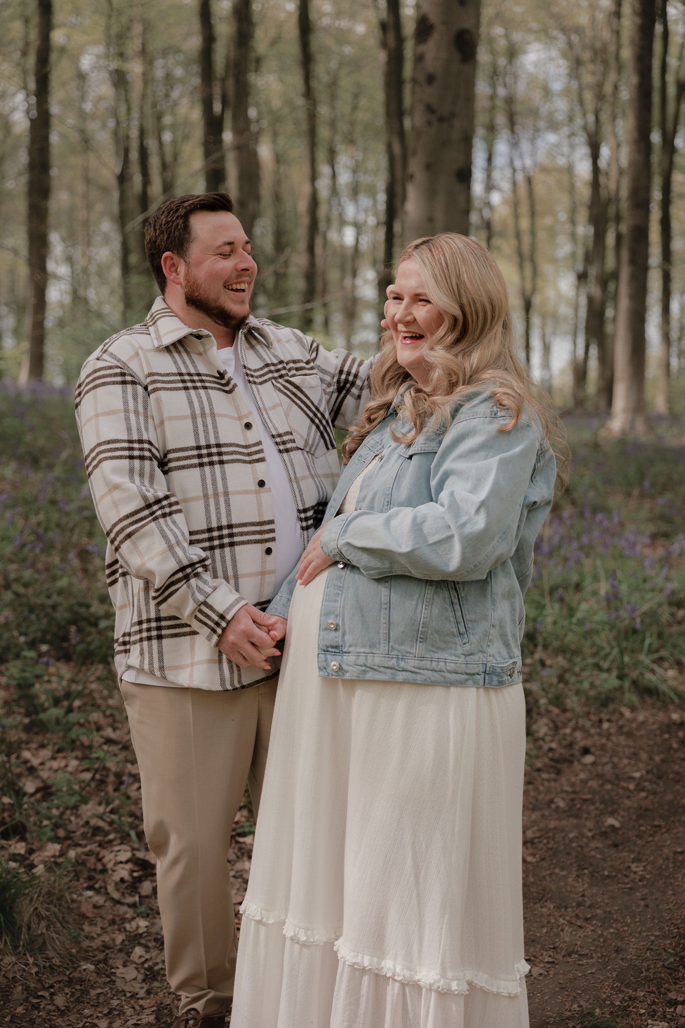A smiling couple poses in a bluebell forest, woman in denim jacket and white skirt, man in plaid shirt and tan pants.