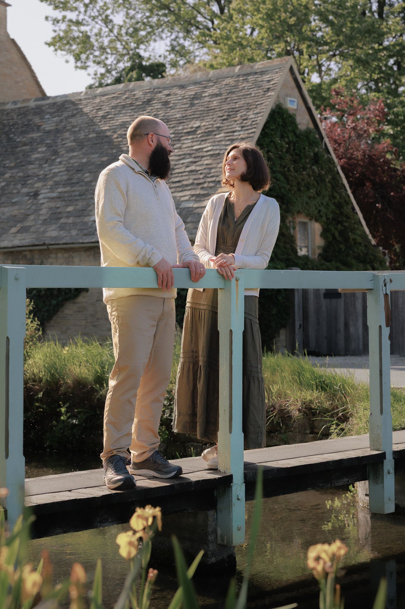 Couple chatting on a wooden bridge beside an ivy-covered stone cottage near Upper Slaughter.