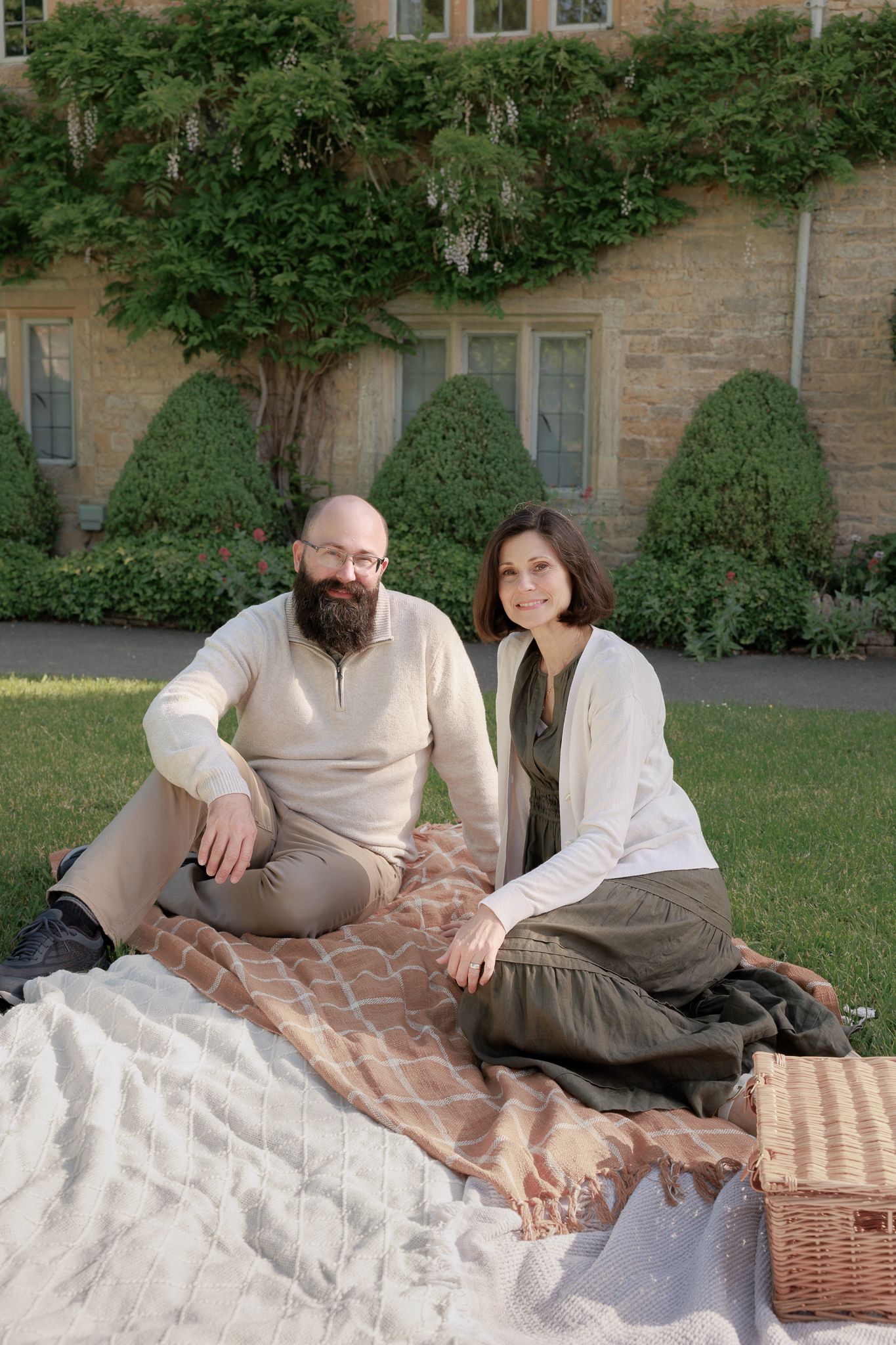 A couple sits on a blanket for a picnic on green grass in front of an ivy-covered stone building in Cotswold, UK.