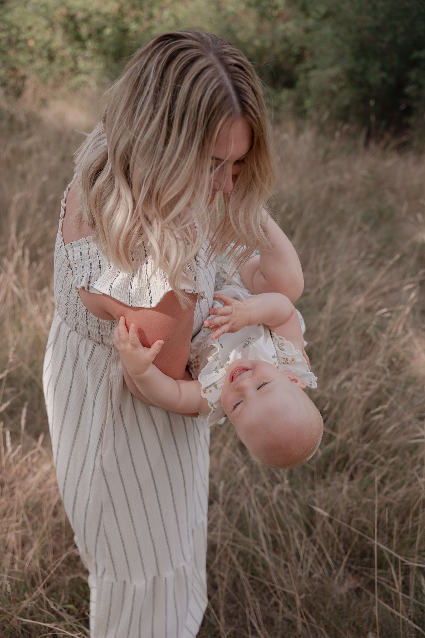 A blonde mother in a striped dress holds her smiling baby upside down in a golden field.